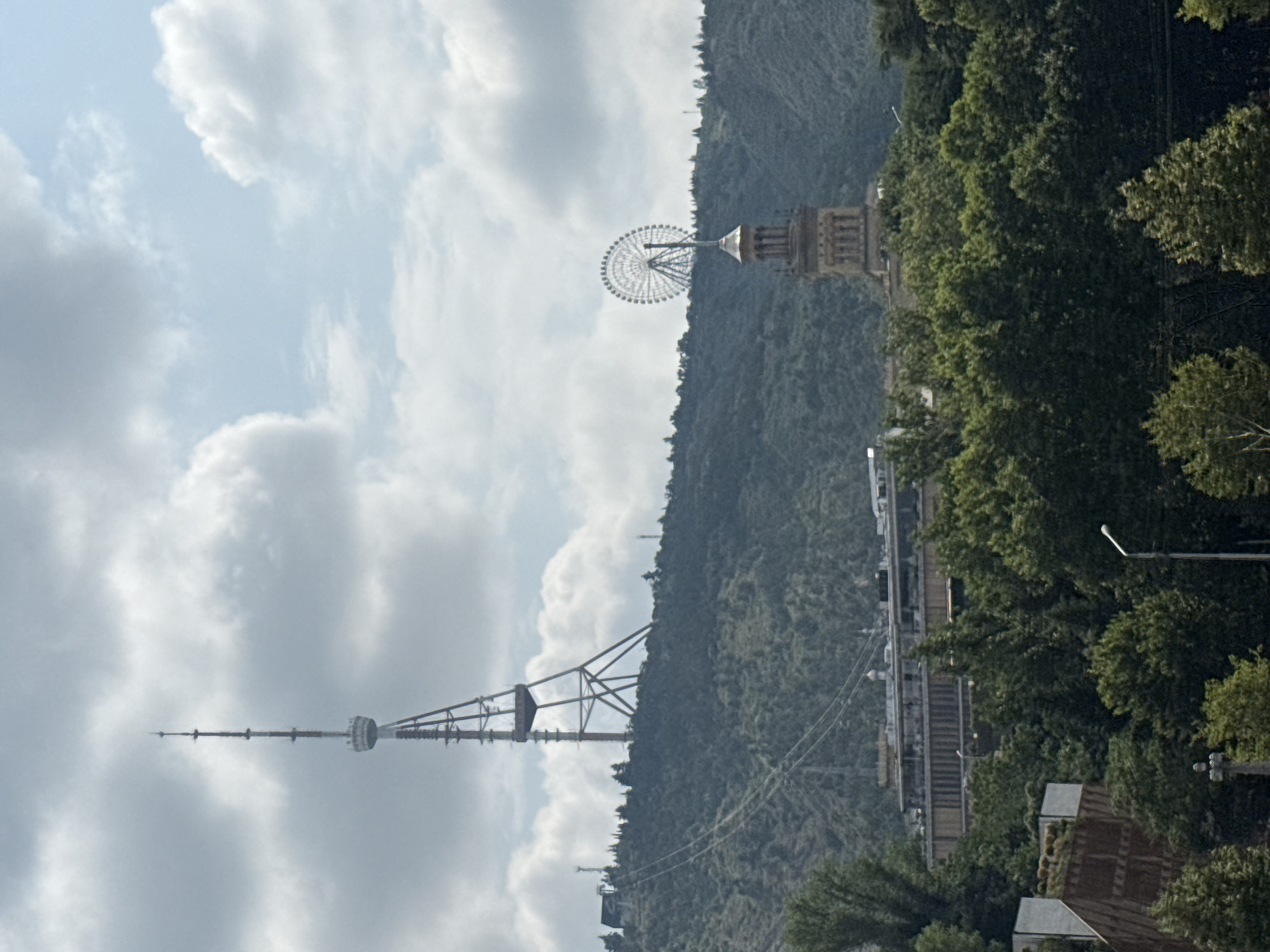 The image captures a panoramic view of the city of Tbilisi, Georgia, dominated by the Mtatsminda Park's attractions. On the left, the prominent Mtatsminda TV Tower rises sharply against a cloudy sky, its lattice structure clearly visible. The cable car lines leading up to the park are also discernible on the green, tree-covered slopes. To the right, a Ferris wheel, a signature attraction of Mtatsminda Park, is perched atop a historic tower, its circular form a striking contrast to the natural landscape. In the mid-ground, a long, multi-story building is situated, partially obscured by dense foliage. The foreground is filled with lush green trees, creating a vibrant natural buffer between the viewer and the cityscape. The overall scene suggests a bright, possibly overcast day, with the prominent landmarks of Tbilisi clearly depicted against a backdrop of rolling hills and a dynamic sky.