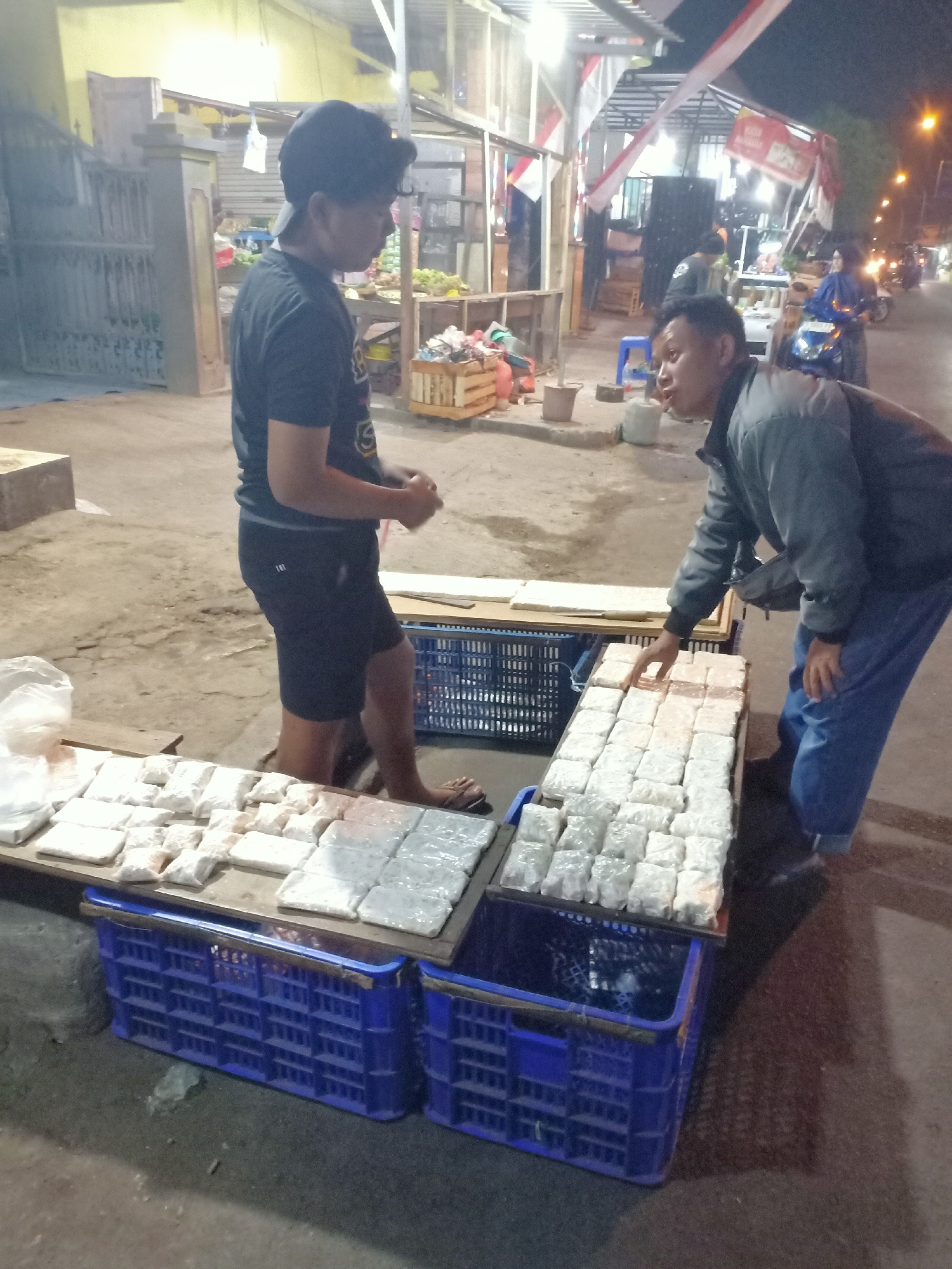 At night, on a street in Semanding, Indonesia, two men stand near a food stall. One man, wearing a black t-shirt and shorts, stands to the left, facing the stall. The other man, wearing a grey jacket and blue jeans, is on the right, leaning over the stall and touching some of the items for sale. The stall displays numerous food items, wrapped in white paper, neatly arranged on wooden planks. Below the planks, blue plastic crates are stacked. In the background, various stalls are visible, illuminated by artificial lights. The street appears to be paved, and there are some vehicles parked in the distance. The overall atmosphere suggests a bustling night market.