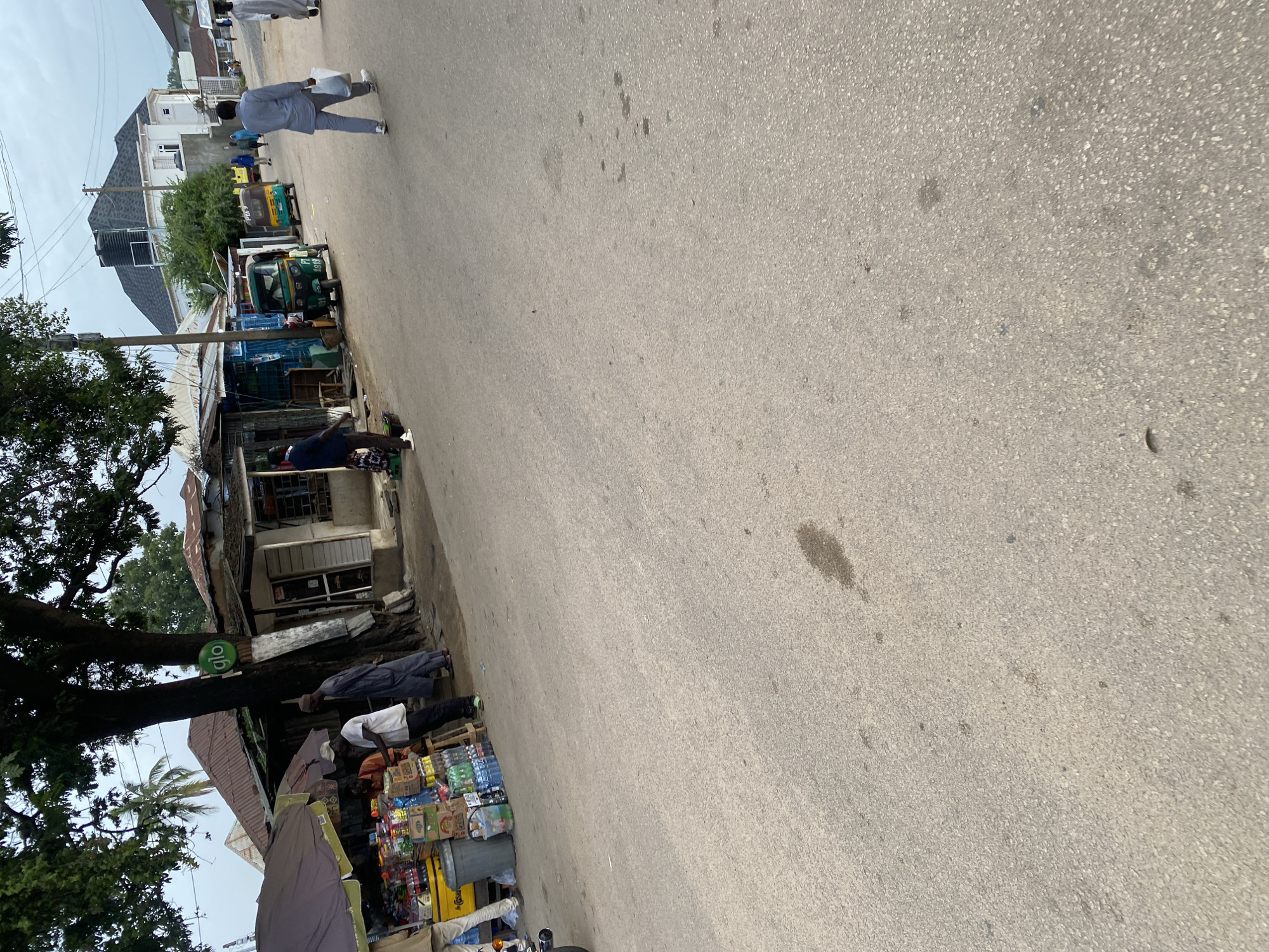 The image shows a street scene in Jos, Nigeria. The foreground is dominated by a paved road with some dark stains. On the left side of the road, there is a stall with various goods displayed, including bottled drinks and other packaged items. Several people are present around the stall, some appearing to be vendors and customers. A large tree with a "glo" sign on it is situated behind the stall. Further down the street, there are more stalls and buildings. Several tricycles, commonly used as public transport in Nigeria, are visible on the road. In the background, a modern building with a dark roof stands out among other structures. People are walking on the road, and some are carrying bags. The overall atmosphere suggests a busy urban environment.