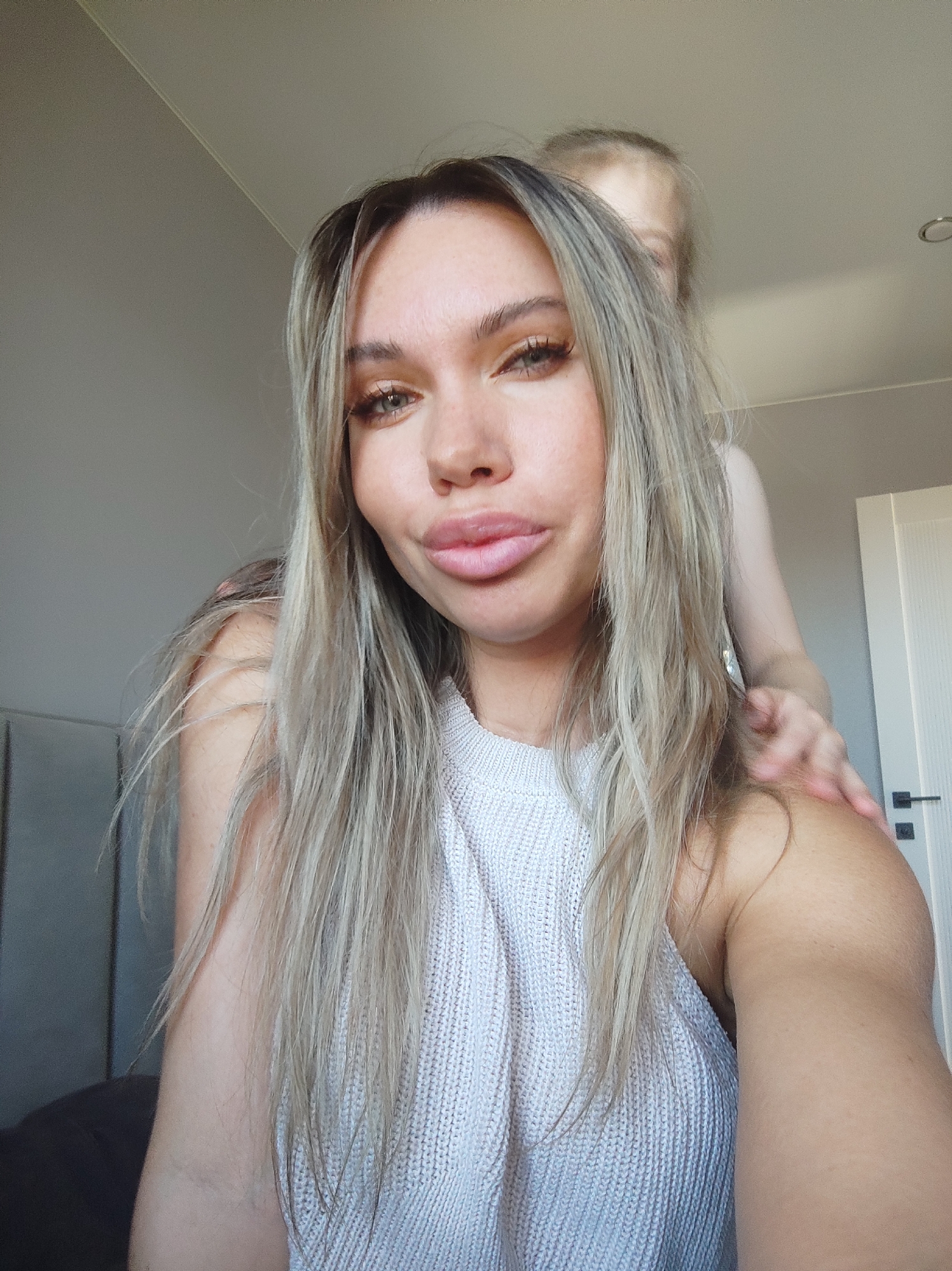 A woman with long, blonde hair and full lips takes a selfie indoors. She wears a white sleeveless top. A child with blonde hair stands behind her, their hand resting on the woman's shoulder. The background includes a light gray wall, a door, and part of a headboard. The scene takes place indoors, in a room with muted lighting.