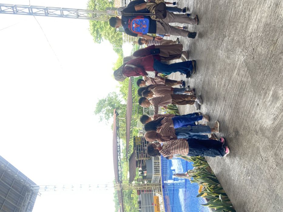 A group of children wearing striped shirts and jeans stand in a line outdoors, near a blue pool. Adults are also present, some with backpacks. The children are facing a structure or building in the background, possibly engaged in an activity or presentation. The scene is bathed in sunlight, with a concrete surface in the foreground.