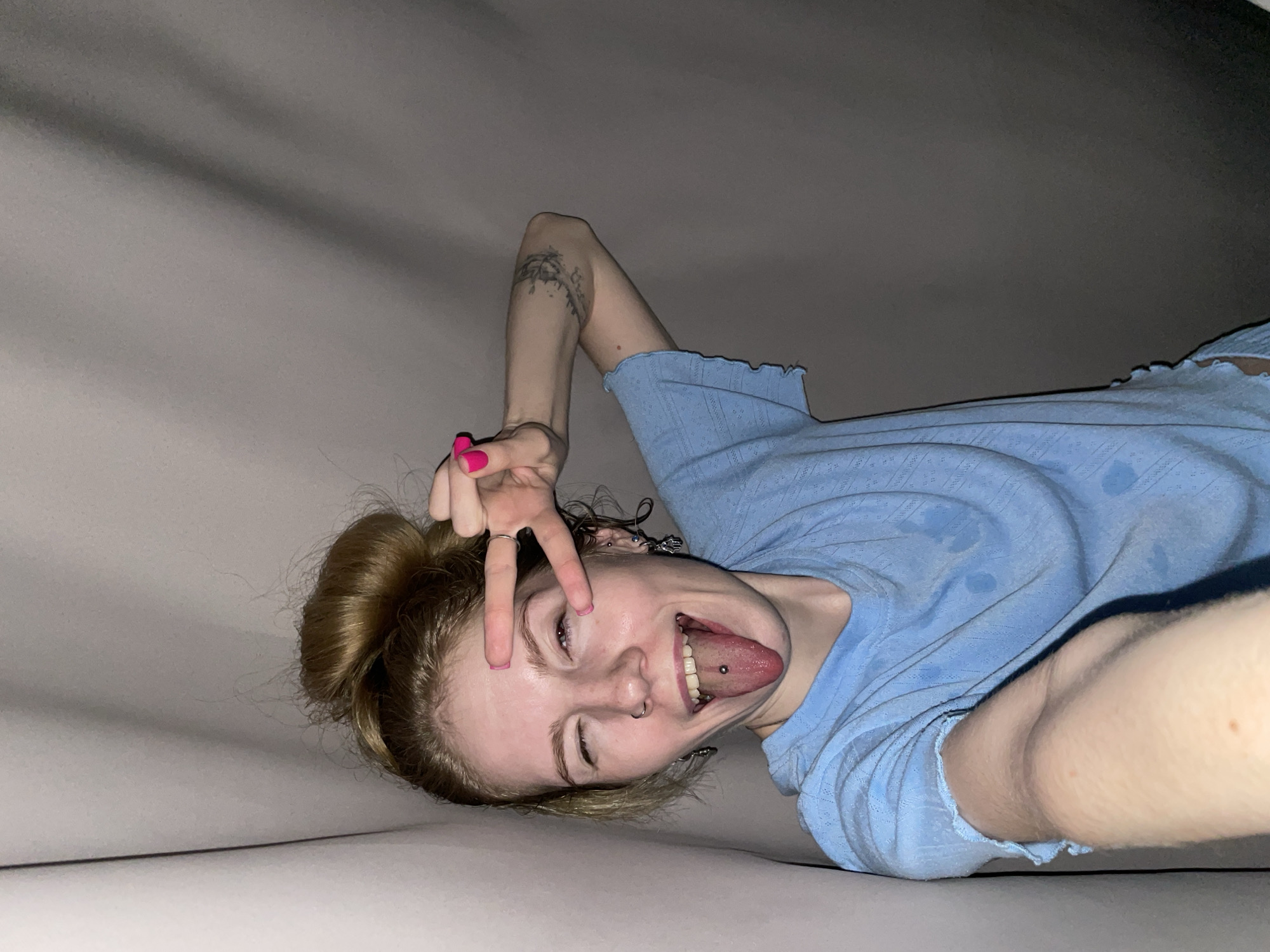 A woman is lying down on a gray surface, possibly a bed. She is wearing a light blue top and has her hair in a bun. She is making a peace sign with her hand, sticking out her tongue, and winking with one eye. She has a tongue piercing and several piercings in her ears. She also has a tattoo on her arm. The location is not discernible from the image.