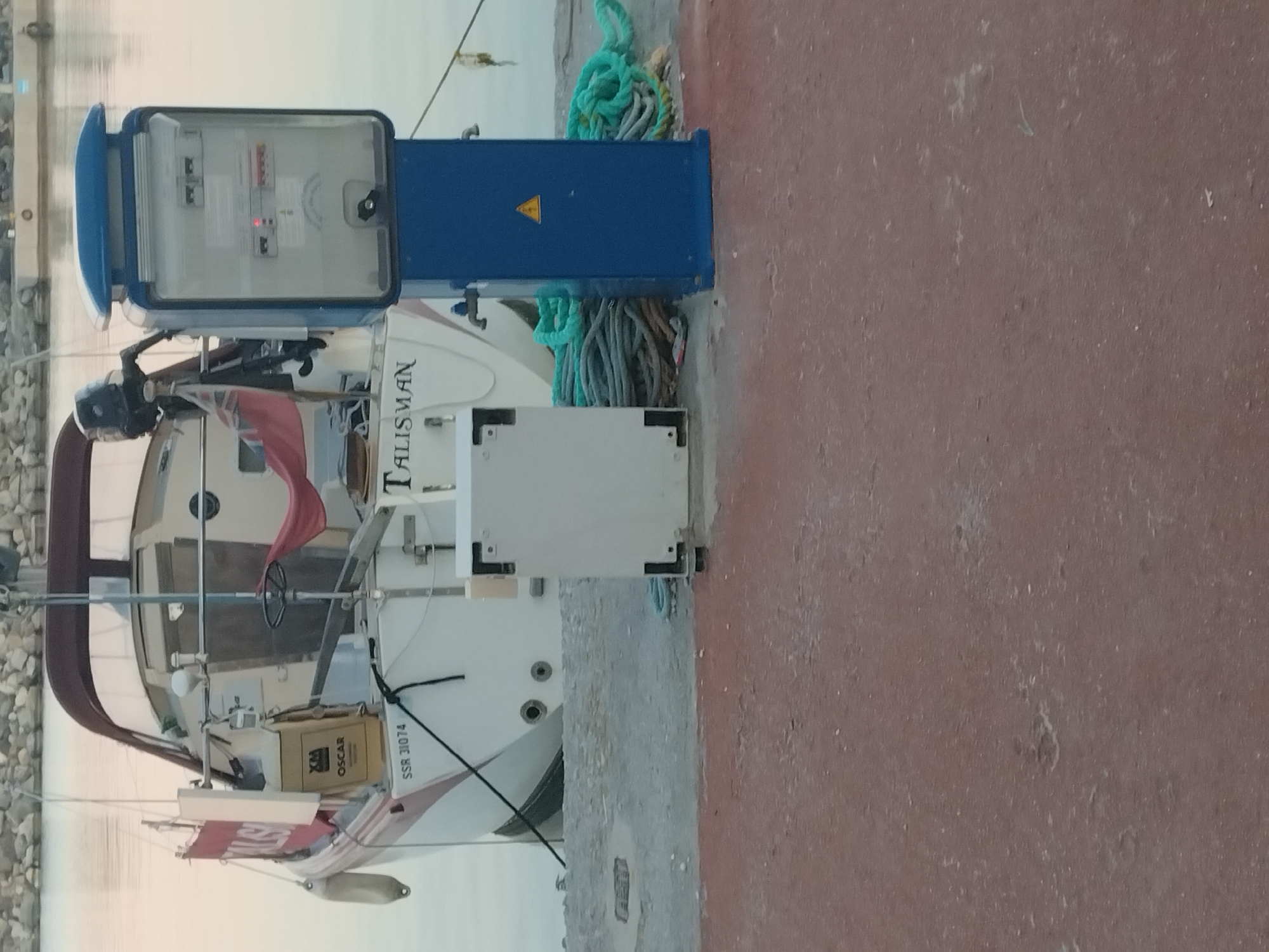 The image shows a boat named "TALISMAN" docked at a marina in Καρδάμαινα, Greece. The boat is white with a maroon canopy. A blue electrical box is adjacent to the boat on the pier. Ropes are also visible near the electrical box. The pier is red. The background shows the waterfront.