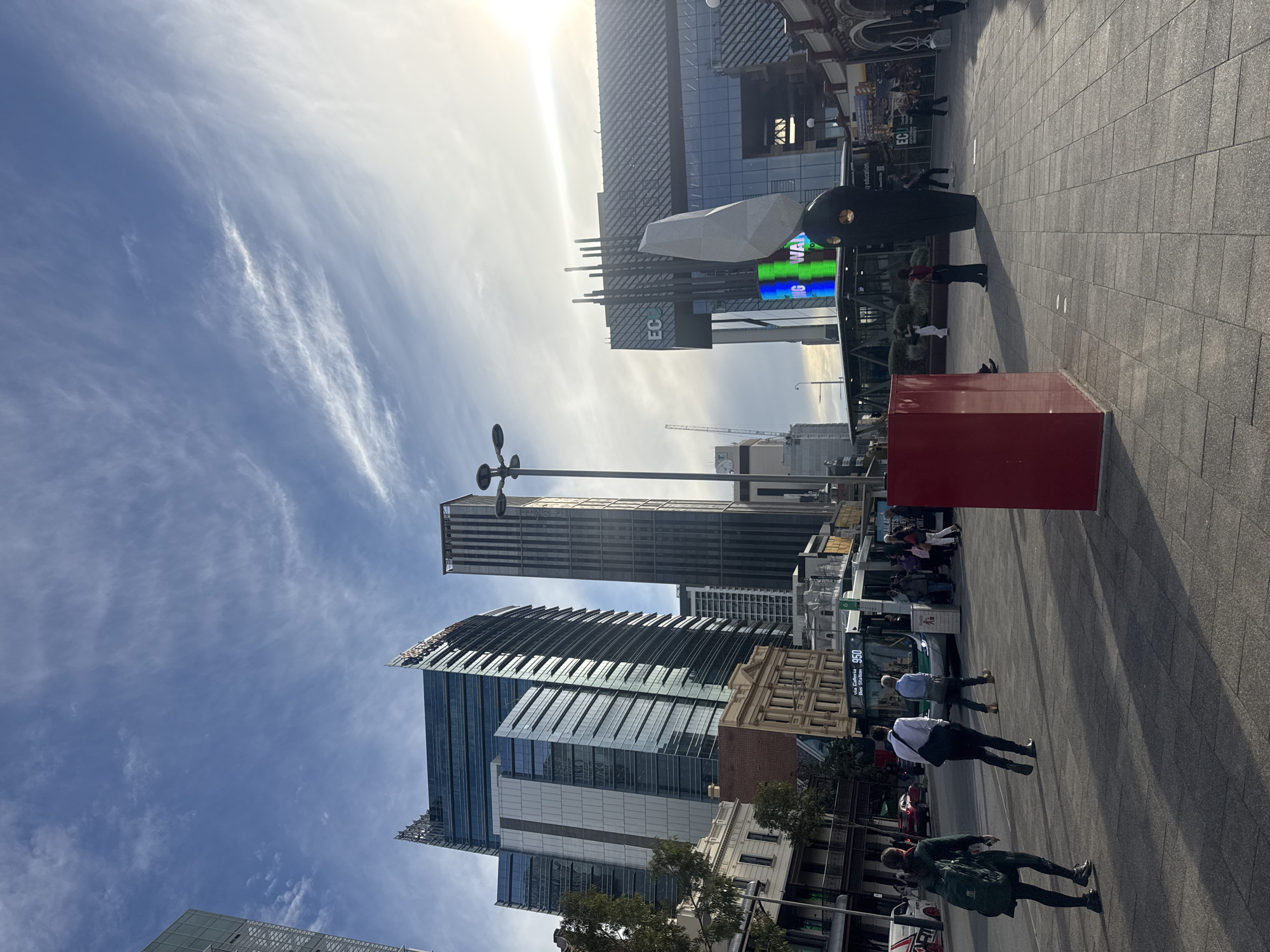 This image depicts a street scene in Perth, Australia. In the foreground, pedestrians are walking on a paved sidewalk, with a bus stop visible nearby. A red cubic sculpture stands prominently on the sidewalk. Tall modern buildings dominate the background, interspersed with some older architectural styles. The sky is blue with scattered clouds and sunlight. The lighting suggests it is daytime. A crane can be seen in the distance, indicating ongoing construction. A large electronic billboard displays colored images and text.