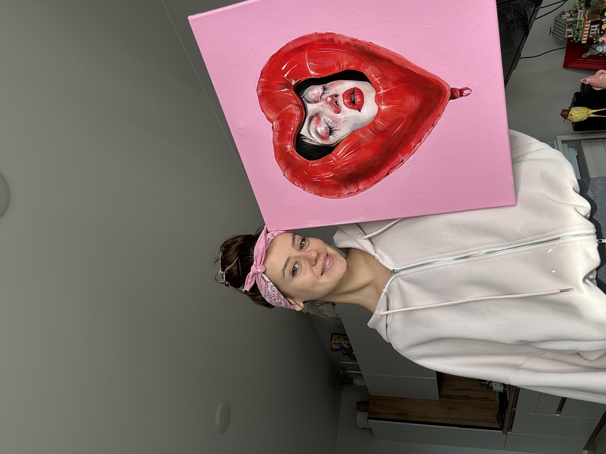 This image, rotated 90 degrees counter-clockwise, depicts a young woman inside a room, likely in Krasnoyarsk, Russia, proudly showcasing a painting. The woman, positioned in the lower right of the frame, smiles warmly at the viewer. She has light brown hair tied back with a pink bandana featuring a white pattern and wears a light pink zippered hoodie.
She holds a square canvas painting that partially covers her head. The artwork features a stylized depiction of a person's face with closed eyes, full red lips, and subtle freckles, framed by a glossy, vibrant red heart-shaped balloon. The background of the painting is a solid, bright pink.
The setting appears to be an indoor space, possibly a studio or living area. In the background, visible to the right of the woman, are various items including what looks like a black computer monitor or television, and shelves displaying numerous small objects, such as miniature structures or figurines, including a distinct yellow bird-like item with a hat. Part of a white cabinet with a wooden top is also visible further down. The wall to the left is a neutral gray, with a circular fixture mounted on it. The lighting is soft and even, suggesting an indoor shot during the day or under artificial light. The woman's joyful expression indicates pride and satisfaction in her artistic creation. No visible text is present.