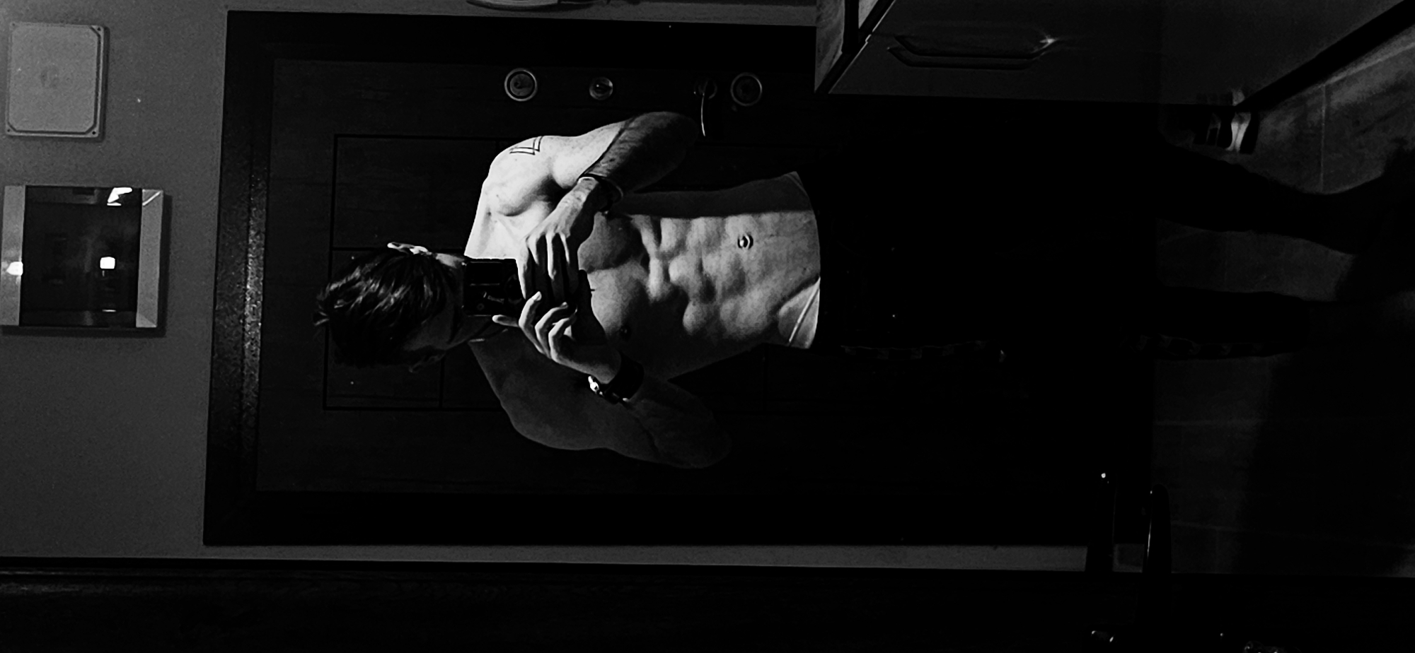 A shirtless man is taking a photo of himself in a mirror. The room appears to be a bathroom, with a mirror and a cabinet visible. There is a small square object and another mirror on the wall adjacent to the larger mirror. The photograph's black and white tone suggests a focus on the man's physique and the interplay of light and shadow. The location is not discernible from the image.