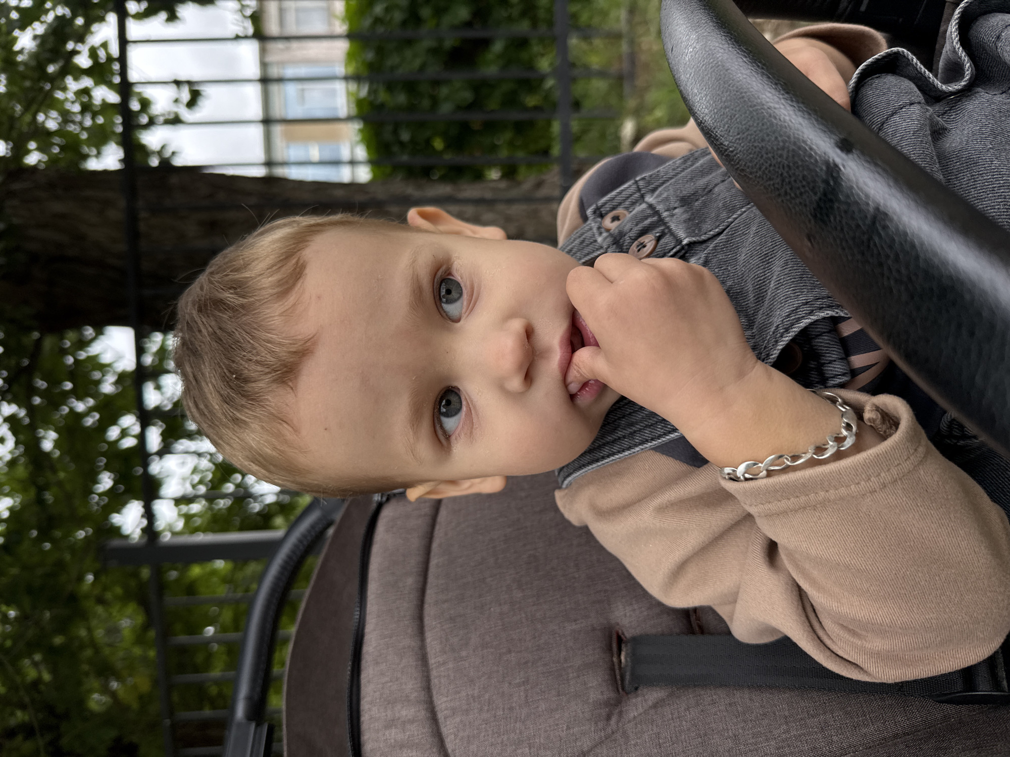 A young child with blonde hair and blue-grey eyes is seated in a stroller, positioned outdoors. The child is wearing a tan long-sleeved shirt and dark grey denim overalls, adorned with a silver bracelet on their left wrist. Their right hand is raised to their mouth, with fingers between their lips. The stroller is a muted brown color with black accents. In the background, a black metal fence encloses greenery and a tree with dappled sunlight filtering through its leaves. Beyond the fence, a building with windows is visible. The overall scene suggests a casual outing in a park or residential area, possibly in the city of Rusanovo, Russia.