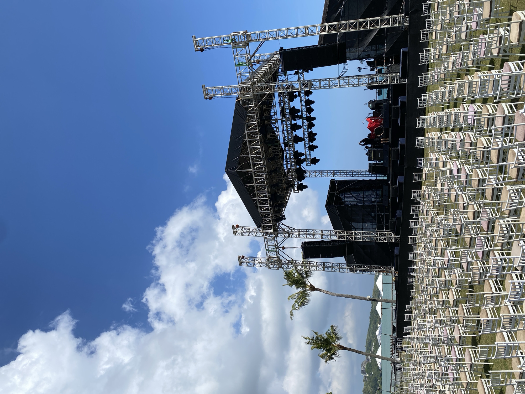 The vertically oriented image captures an outdoor event venue, likely a concert or festival setup, under a bright blue sky with scattered white clouds. A large stage structure made of silver metal trussing and covered with black tarpaulin dominates the scene. Numerous stage lights are mounted on the truss, along with large sound speaker systems, including hanging line arrays and ground-stacked speakers.
In front of the stage, hundreds of white chairs with light-colored seats are meticulously arranged in long, orderly rows on a grassy field, indicating preparation for a large audience. On the stage itself, parts of a drum set are visible, and two individuals, likely crew or technical staff, are present; one in dark clothing is near equipment on the right side of the stage, and another in red is partially obscured.
Two tall palm trees stand in the foreground on the lower right, adding to the tropical ambiance. Beyond the seating area, a calm body of water, likely the ocean, is visible, with a distant landmass or hills forming the horizon. The scene is bathed in bright daylight, suggesting fair weather during late morning or early afternoon. This setting, with its stage, seating, palm trees, and ocean backdrop, points to an outdoor performance or gathering venue in a coastal or tropical location, aligning with Kuta Selatan, Indonesia. No visible text is present in the image.
