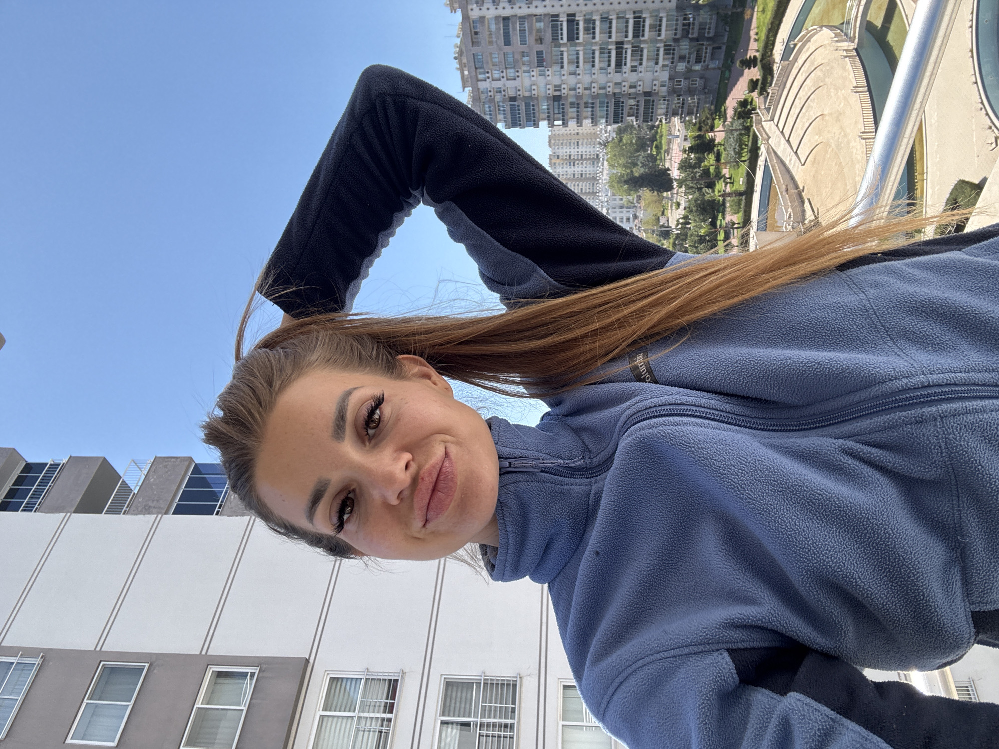 A young woman wearing a blue and black fleece jacket is taking a selfie outdoors. She is leaning over a railing, with city buildings visible in the background. Her hair is pulled back in a ponytail. In the background are buildings and what appears to be a park or garden with a fountain. The location is Erdemli, Turkey, based on architectural styles.
