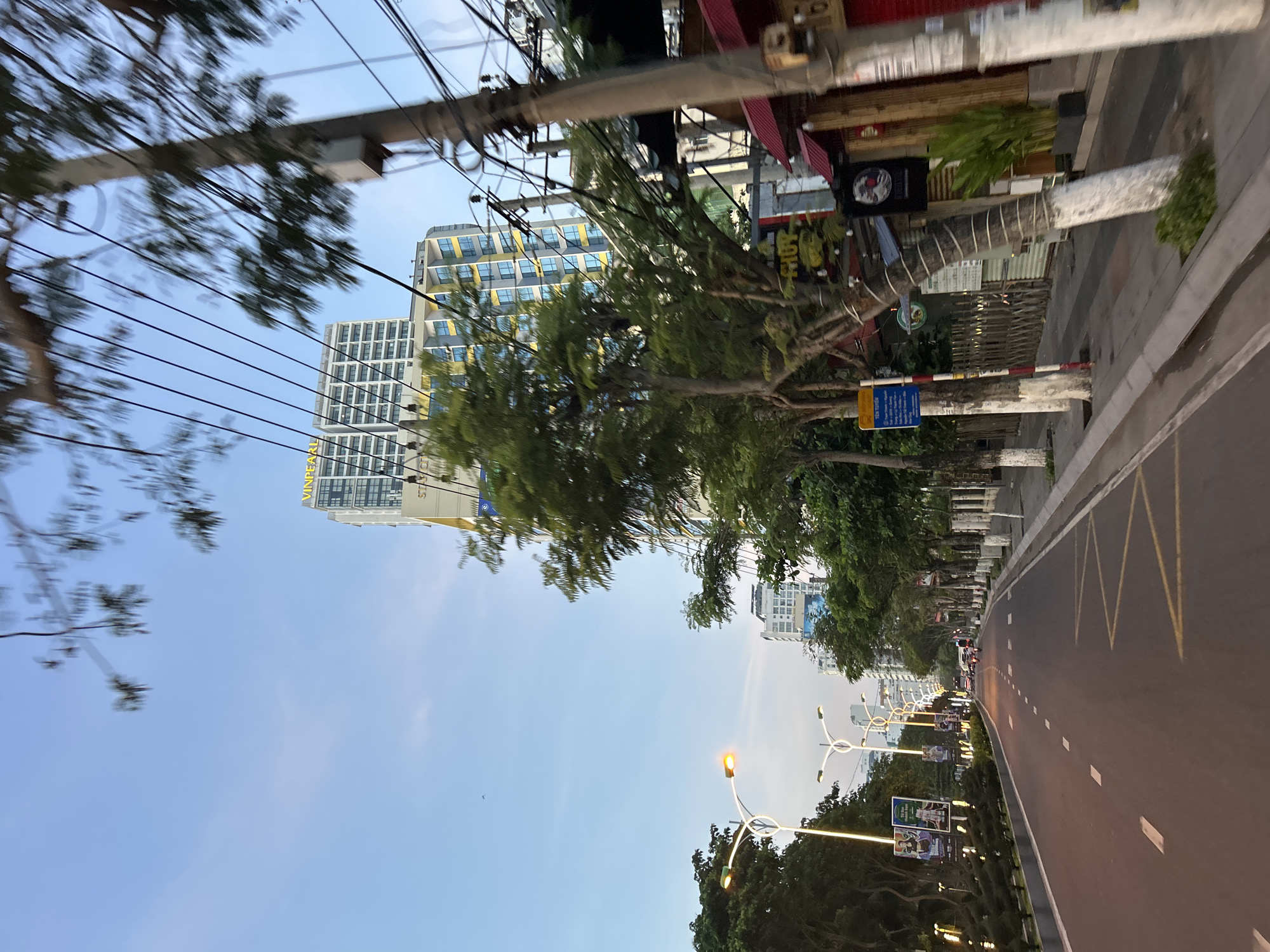 The image captures a street scene in Nha Trang Ward, Vietnam, during what appears to be either early morning or late evening due to the low light and illuminated street lamps. The perspective is from a moving vehicle, as indicated by the motion blur in the foreground and on the trees.
The street itself is paved with asphalt and marked with white dashed lines indicating lanes and yellow zig-zag markings for a no-parking zone. On the left side of the street, there is a landscaped median with trees and decorative bushes, and several modern, arching streetlights are illuminated. Billboards are also visible along this side.
On the right side of the street, there is a sidewalk lined with trees whose trunks are painted with white and red bands. Beyond the sidewalk, several buildings are visible. The most prominent is a tall, modern high-rise building with a "VINPEARL" sign near its peak. In front of it, another multi-story building with yellow and blue accents can be seen. Further down the street, other buildings and structures suggest a commercial or mixed-use area, with signage for businesses, including what appears to be a Japanese restaurant with red awnings and a black sign featuring a circular emblem. Electrical poles with numerous wires are also present along the sidewalk.
The sky is a pale blue with some wispy clouds. A few vehicles, including motorcycles and a bus, are present on the road in the distance. There are no clearly identifiable individuals captured in detail due to the motion blur and distance. The overall impression is of a vibrant urban environment.