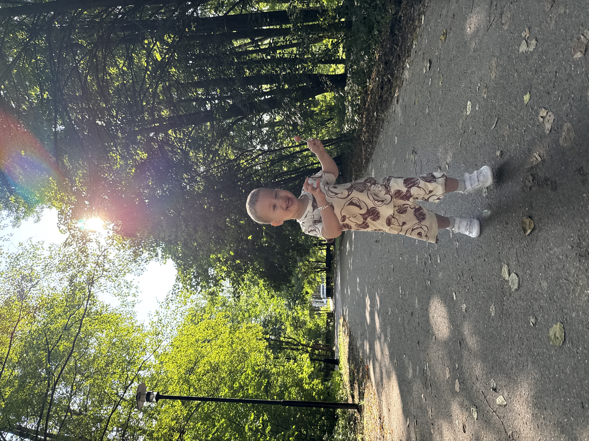 A young child with blonde hair and a light-colored t-shirt with a Mickey Mouse pattern is walking down a paved path in a park-like setting. The child is smiling and holding a pink lollipop in their right hand. The path is surrounded by trees with green leaves, and the sun is shining through the canopy, creating dappled shadows on the ground. There is a lamppost on the left side of the path, and a car is visible in the distance on the right. The overall scene depicts a bright, sunny day in a natural environment. The location is specified as Rusanovo, Russia.