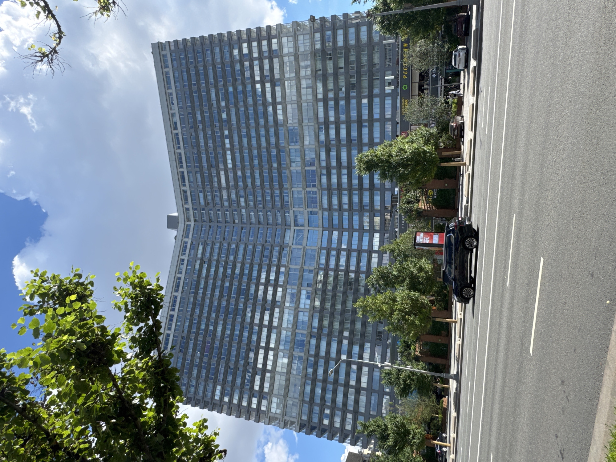 The image shows a large, curved building with a facade of many windows, situated in Moscow, Russia. The building appears to be an apartment complex or hotel. In front of the building is a wide street with a multi-lane road, where several cars are visible. There are trees lining the sidewalk in front of the building, and a lamppost stands near the road. The sky is blue with some white clouds, and the sun is shining, casting shadows. The overall scene is an urban environment on a clear day.