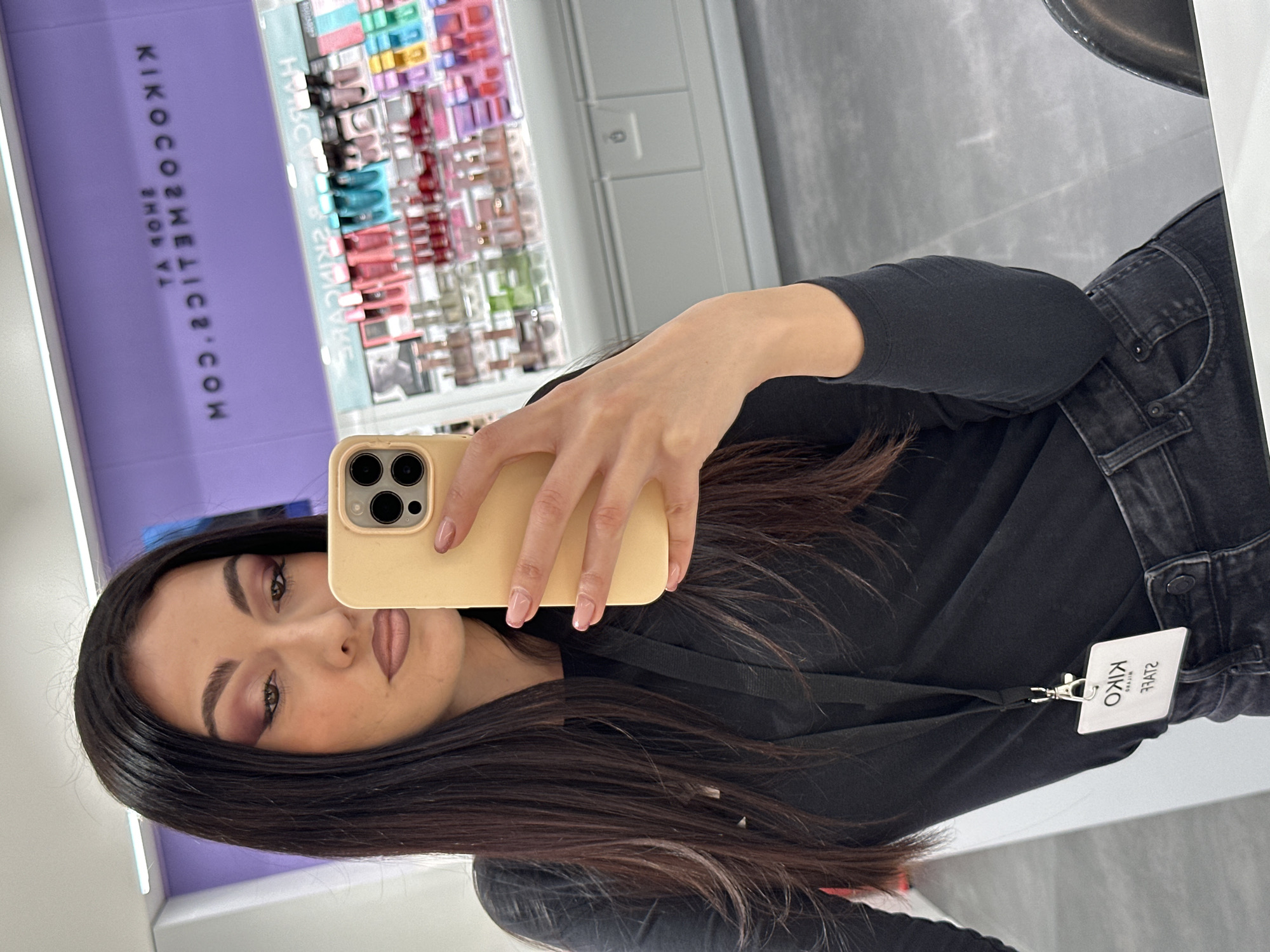 A woman is captured in a mirror selfie, positioned upright as if taking the photo herself. She has long, dark hair that cascades over her shoulders and is wearing a black long-sleeved top and dark-colored jeans. Her left hand holds a light yellow smartphone, which features multiple camera lenses, up to take the picture. Her fingernails are painted with a light pink or nude polish.
Her makeup is visible, featuring defined eyebrows, dark eyeshadow with hints of purple or plum, and a nude lipstick. Attached to her top on the right side is a white ID badge, clearly displaying "KIKO MILANO" and "STAFF" in legible, albeit reflected, text.
The background, a reflection in the mirror, shows the interior of a cosmetics store. A prominent purple sign or wall is visible on the left, with white mirrored text that appears to read "KIKOCOSMETICS.COM" and "SHOP AT" when deciphered. To the right of this sign, shelves are stocked with an array of colorful cosmetic products in various packaging, including shades of pink, red, blue, green, and purple, creating a vibrant display. The setting appears to be a well-lit retail space, indicative of a KIKO MILANO store, where the woman, presumably a staff member, is taking a personal photo.