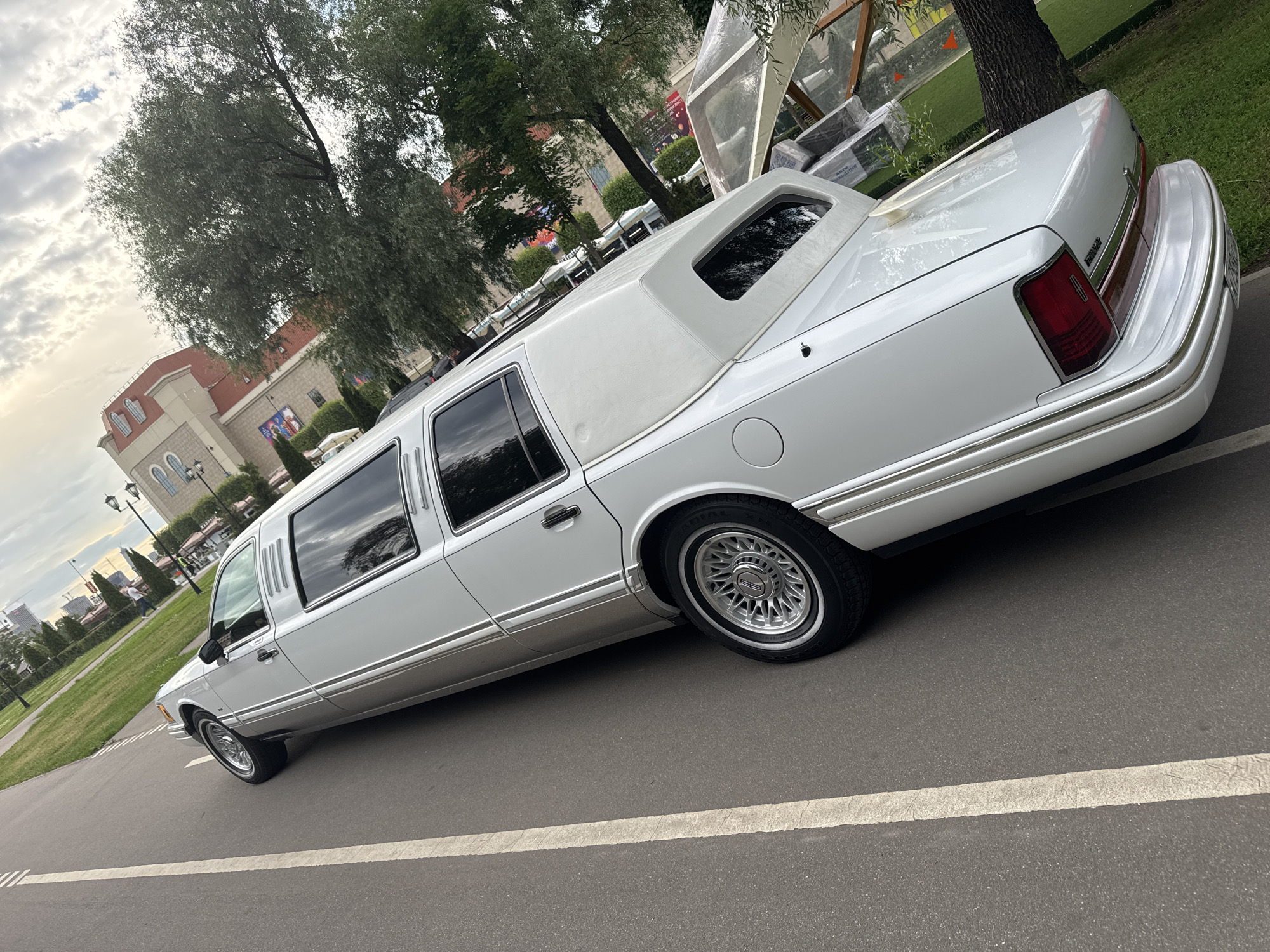 The media file shows a white stretch limousine parked on an asphalt road in what appears to be a park or public area. The limousine is the primary subject, seen from its rear quarter, extending into the foreground. It has a white vinyl roof, tinted windows, and chrome trim along its body. The wheels have a distinctive multi-spoke design with a chrome finish.
In the background, several large trees with lush green foliage dominate the scene. Behind the trees, there's a glimpse of a building with a red roof and arched windows, suggesting an architectural style common in urban European settings. There are also elements that suggest an outdoor seating area with tables and chairs, possibly a cafe or restaurant. The sky is partly cloudy, indicating a daytime scene, likely late afternoon or early evening given the lighting.
There is no visible activity of people in the immediate vicinity of the limousine. The overall impression is one of stillness, with the limousine appearing to be either waiting or recently parked. The location context provided states Moscow, Russia, which aligns with the architectural style and the overall urban park setting. There is no visible text in the media file.
