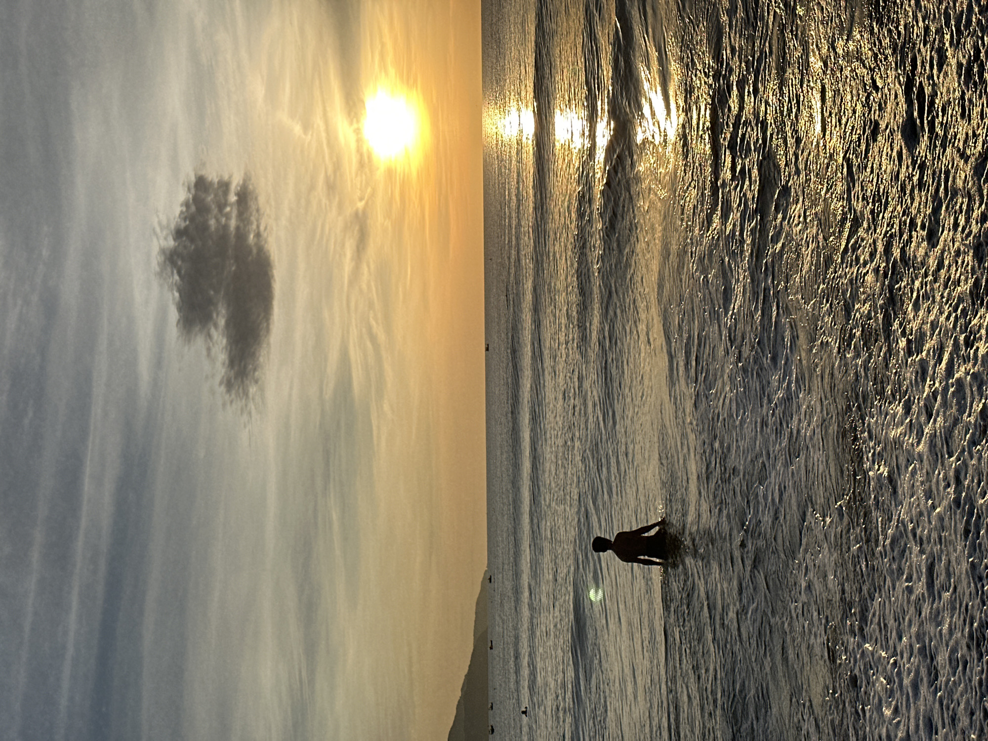 A solitary, silhouetted figure stands waist-deep in the ocean, facing away from the viewer, towards the sunrise over the sea. The sun, a bright orb, casts a shimmering golden path across the water's surface. The sky is a gradient of soft blues and oranges, with wispy clouds and a single, darker cloud in the upper left quadrant. In the distance, a faint coastline with mountains can be seen, dotted with several small boats. The foreground is filled with foamy waves lapping at the shore. The scene is likely captured at sunrise on one of Đà Nẵng, Vietnam's beaches, such as Mỹ Khê or Phạm Văn Đồng Beach.