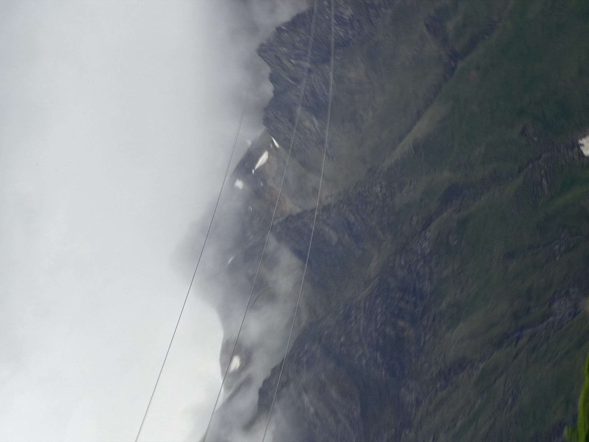 The image shows a mountain landscape shrouded in fog. Several thin wires crisscross the foreground, running horizontally across the frame. The mountains themselves are steep and rocky, with patches of green vegetation on their lower slopes. The sky is overcast and the overall atmosphere is hazy. There are no people or specific structures visible in the scene. The location is unknown.
