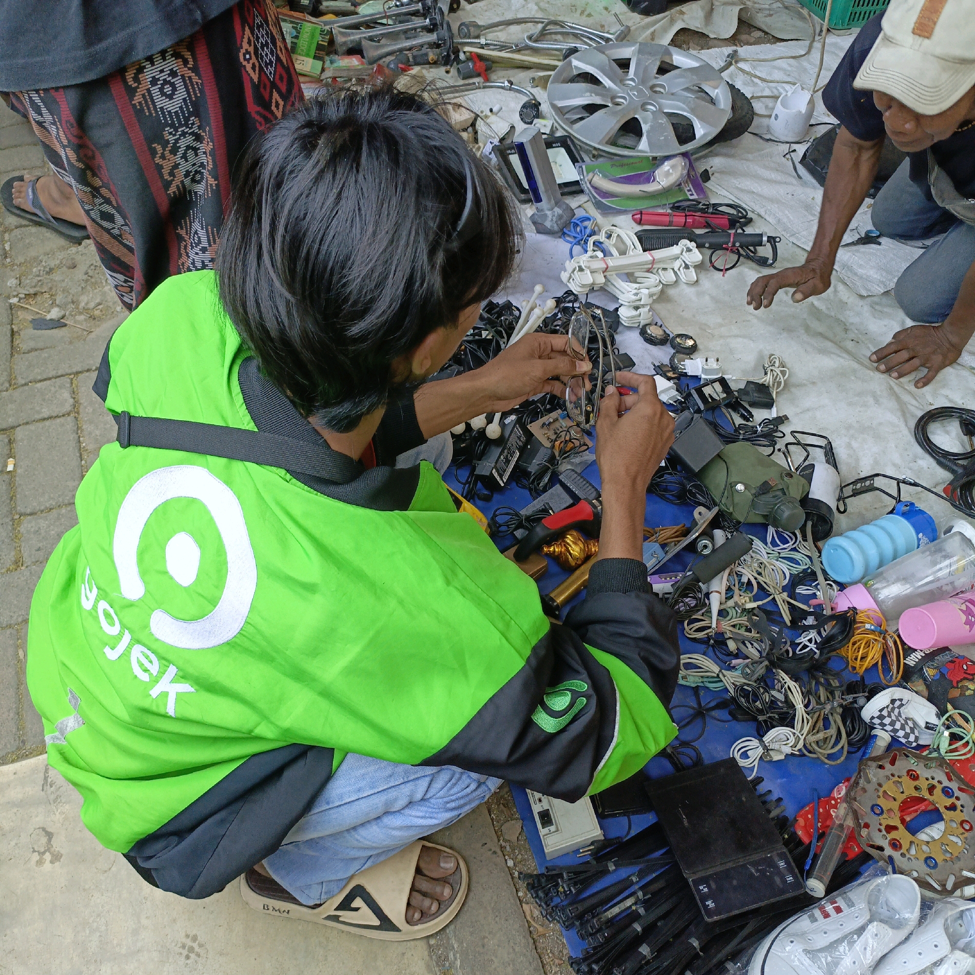 In Ambarawa, Indonesia, a street market scene unfolds on a blue tarp laid out on the ground, displaying a variety of items. Two individuals are present. One, wearing a bright green Gojek jacket, is engrossed in examining a pair of eyeglasses. This person is wearing blue jeans and tan flip-flops with a black triangular logo. The second individual, partially visible, wears a dark t-shirt, blue jeans, and a light-colored baseball cap, and appears to be reaching towards the merchandise. The items for sale include numerous electronic cables, adapters, a digital scale, a water bottle, a car wheel cover, and a motorcycle brake disc. The background suggests an outdoor setting, possibly on a paved surface.