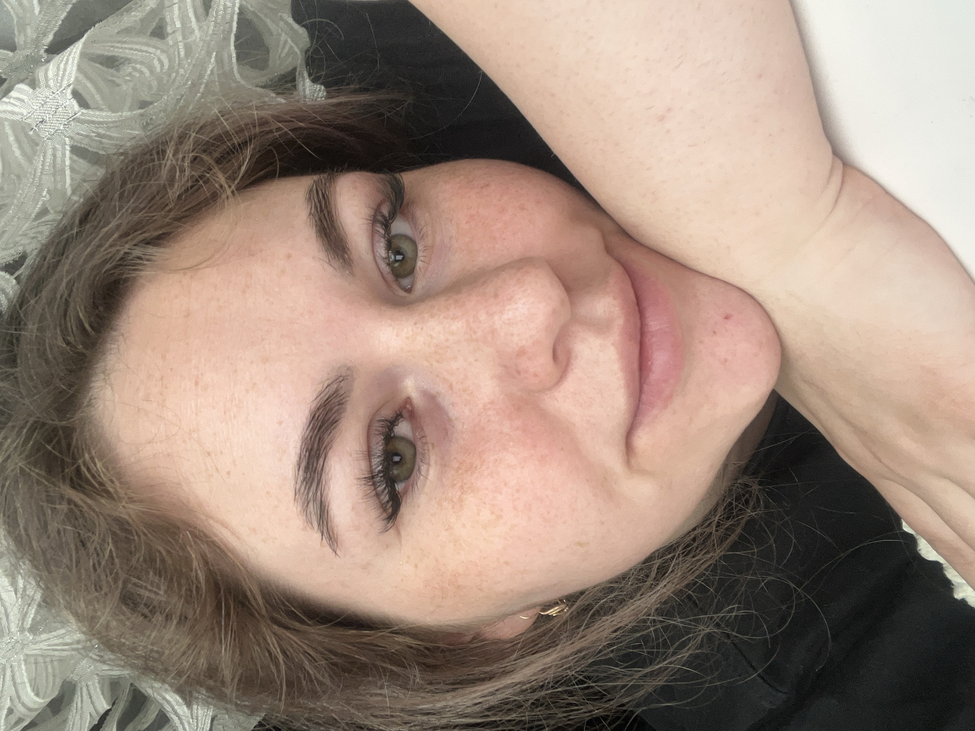 A close-up, eye-level portrait features a young person, likely female, lying down and looking directly at the camera with a relaxed expression and a soft smile. The subject has fair skin with a scattering of freckles across her nose and cheeks. Her eyes are green or hazel, accentuated by long, dark eyelashes and well-defined, dark eyebrows. Her brownish hair, appearing slightly disheveled, frames her face. A small, golden-colored earring is visible in her left earlobe. She is wearing a dark-colored garment, possibly a top, which is partially visible around her neck and shoulder.
The background consists of a light-colored, intricately patterned fabric, possibly a pillow or blanket, on the left side, and a plain white surface on the right. The lighting is soft and even, indicating an indoor setting. The person's pose is casual and comfortable, with one arm visible near her head on the right side of the frame. There is no visible text in the image. This photo was taken in Chaikovsky, Russia.