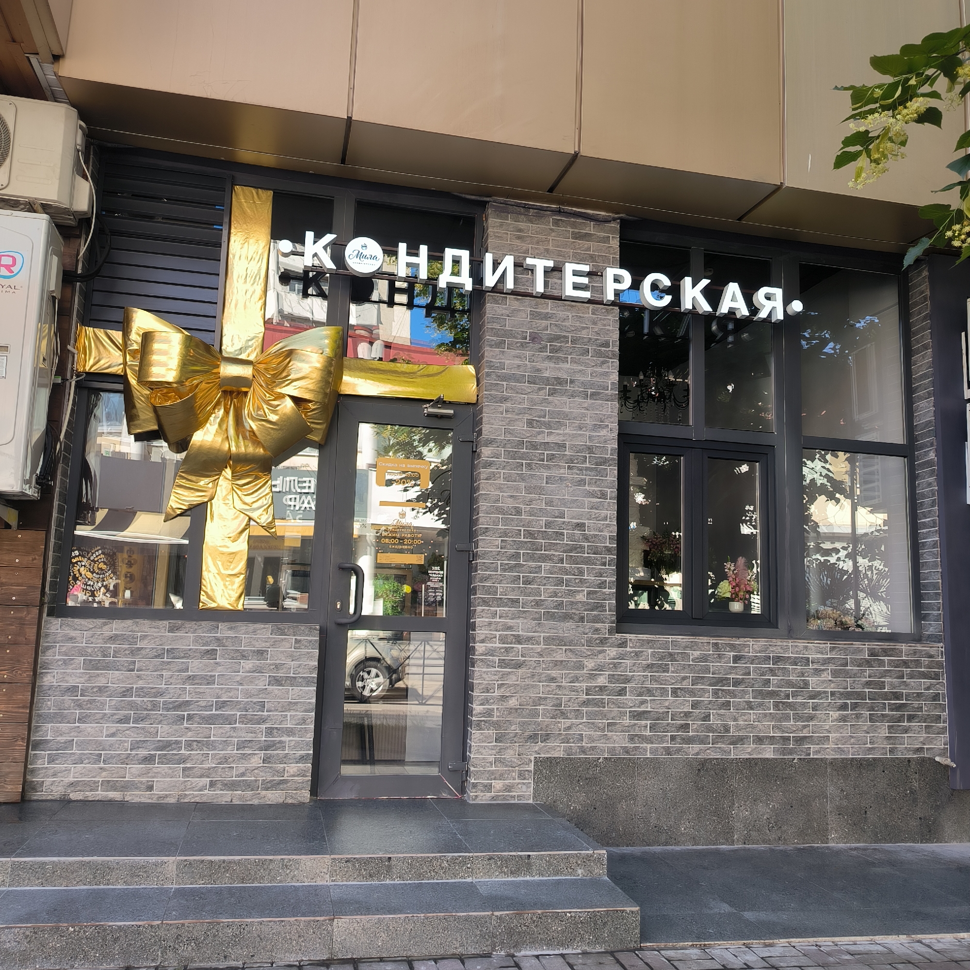 The image shows the storefront of a confectionery shop named "Кондитерская" (Confectionery) in Sochi, Russia. The shop's facade is decorated with a large, shiny golden bow, resembling a gift wrapping. The text "КОНДИТЕРСКАЯ" is prominently displayed in white illuminated letters above the entrance. A smaller circular logo with the word "Мила" is also visible as part of the signage. The entrance consists of a glass door with a golden bow decoration also covering part of it. Inside the shop, reflections show various items, including chandeliers and floral arrangements. The exterior walls are made of grey brick. To the left of the entrance, an air conditioning unit is mounted on the wall. The shop is situated on a street with a paved walkway and steps leading up to the entrance.