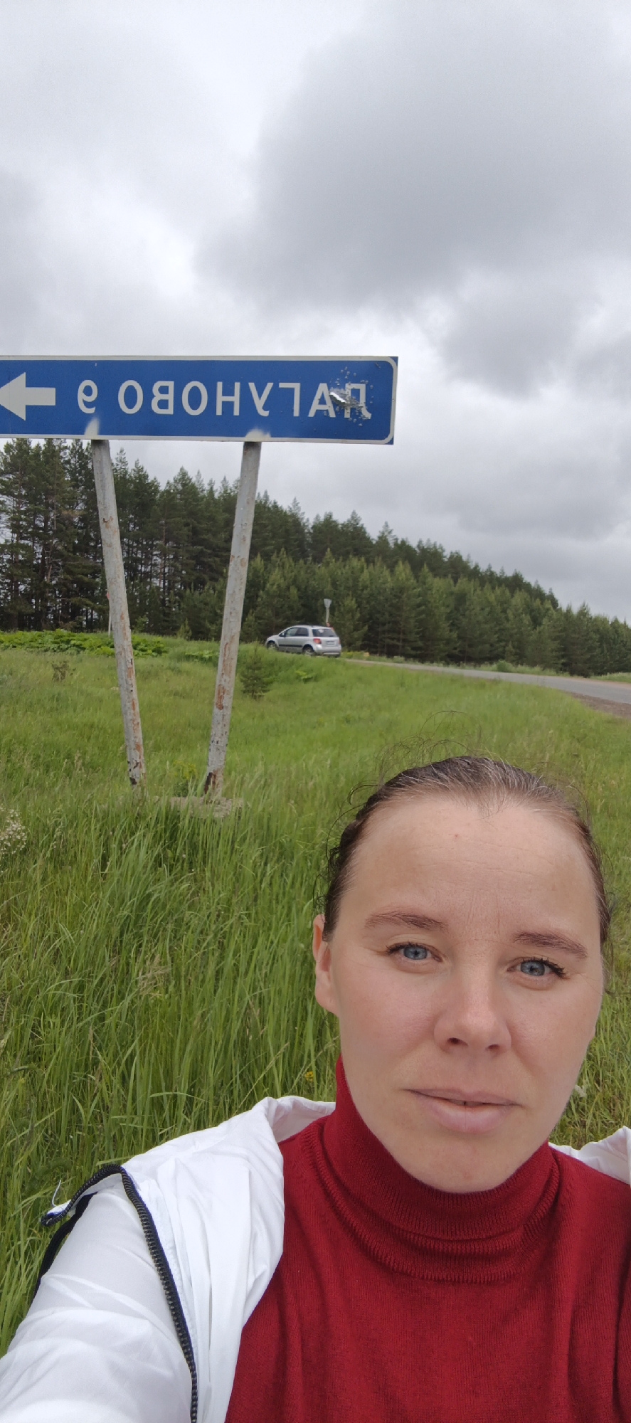 An adult female is depicted in a selfie, positioned in the foreground of a rural outdoor scene in Makshaki, Russia. She has light skin, blue eyes, and light brown hair pulled back. She is wearing a red turtleneck sweater under a white zippered jacket and is looking directly at the camera. Behind her, a blue rectangular road sign with white mirrored Cyrillic text, which appears as "ЯТЛАНУВО О" and includes a left-pointing arrow, is mounted on two weathered metal poles. In the middle ground, a silver SUV is visible on a paved road that curves into a dense evergreen forest. The immediate foreground consists of tall, verdant green grass and other foliage. The sky above is overcast with light gray clouds.