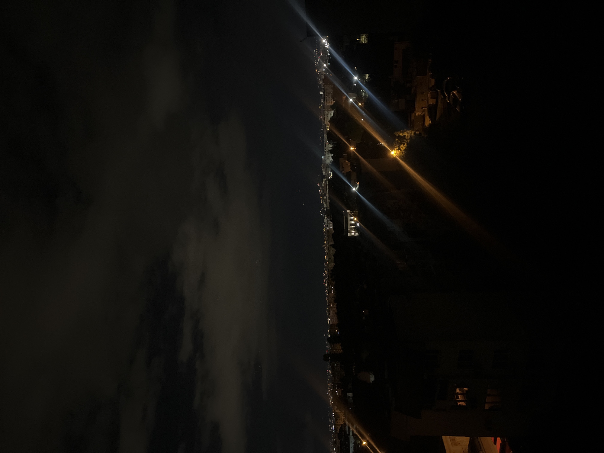 This is a night-time shot of the city of Sofia, Bulgaria. The scene captures a coastline at night, with a sky that appears to be cloudy. A long line of lights stretches across the midground, possibly indicating a road or buildings along the coast. The darkness obscures most of the details, but the lights suggest the presence of urban structures. The right side of the image is dominated by dark shadows and a few brighter areas that could indicate buildings or other structures. The focus seems to be on the city lights and the contrast between the dark night sky and the illuminated cityscape.