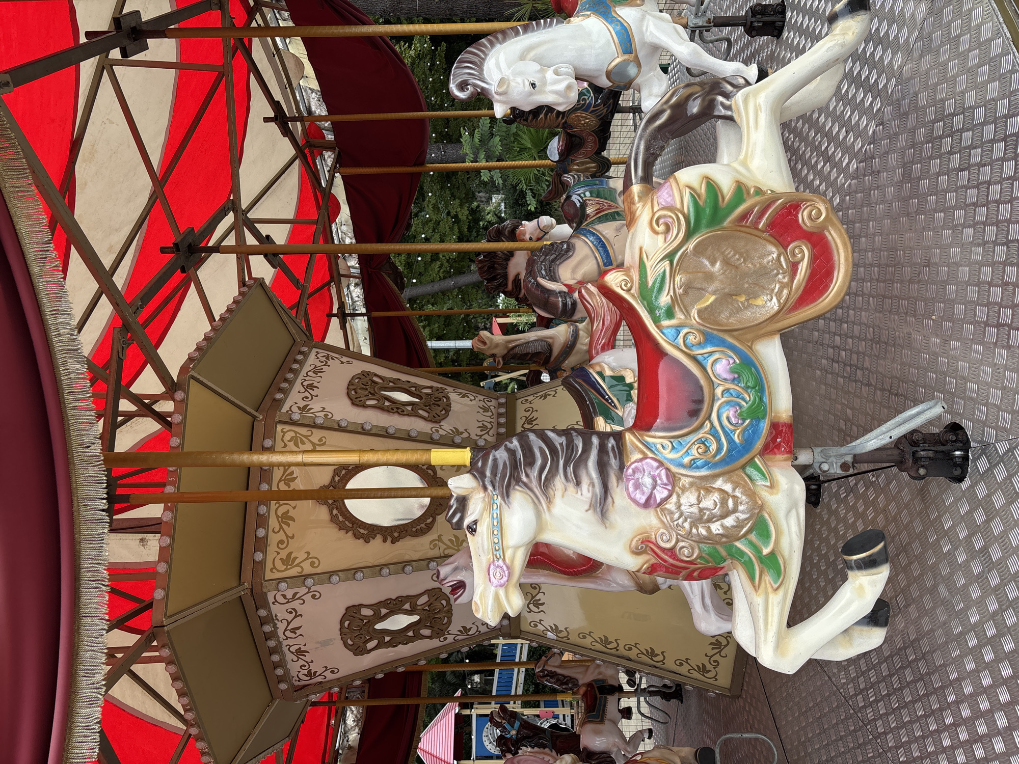 The image shows a close-up of a carousel with several painted horses. The carousel has a red and white striped canopy and is decorated with ornate patterns. In the foreground, a white horse with a brown mane and tail is prominently featured, adorned with a saddle painted in vibrant colors, including blue, red, green, and gold. The saddle features a depiction of a lion's head and an eagle. Behind this horse, other carousel horses are visible, including a white horse with a brown mane and a brown horse. The carousel floor is made of a metallic, textured material. In the background, greenery and trees suggest an outdoor setting. To the left, a glimpse of a children's playground structure with a red roof is visible. The image appears to be taken in Sochi, Russia, judging by the overall aesthetic and common park attractions found in the city.