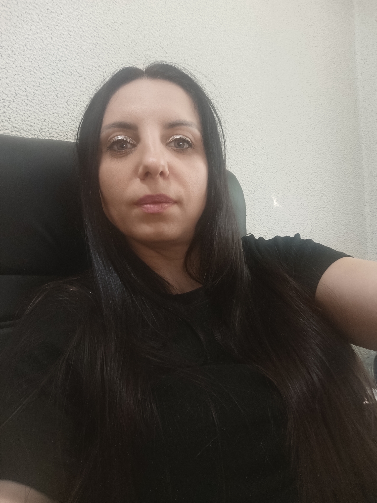 This is a medium shot, eye-level, indoor photograph of a woman with long, dark hair. She is sitting in a black chair, facing the camera. She is wearing a black t-shirt. Her makeup includes eyeshadow with glitter and lip gloss. The background is a textured, light-colored wall. The lighting is somewhat dim, suggesting an indoor setting that is not brightly lit, potentially an office or a room with artificial lighting. There are no other people or objects clearly visible in the frame. The woman's expression is neutral, and there are no strong indications of emotion. No text is visible in the image. The photograph appears to be a selfie.