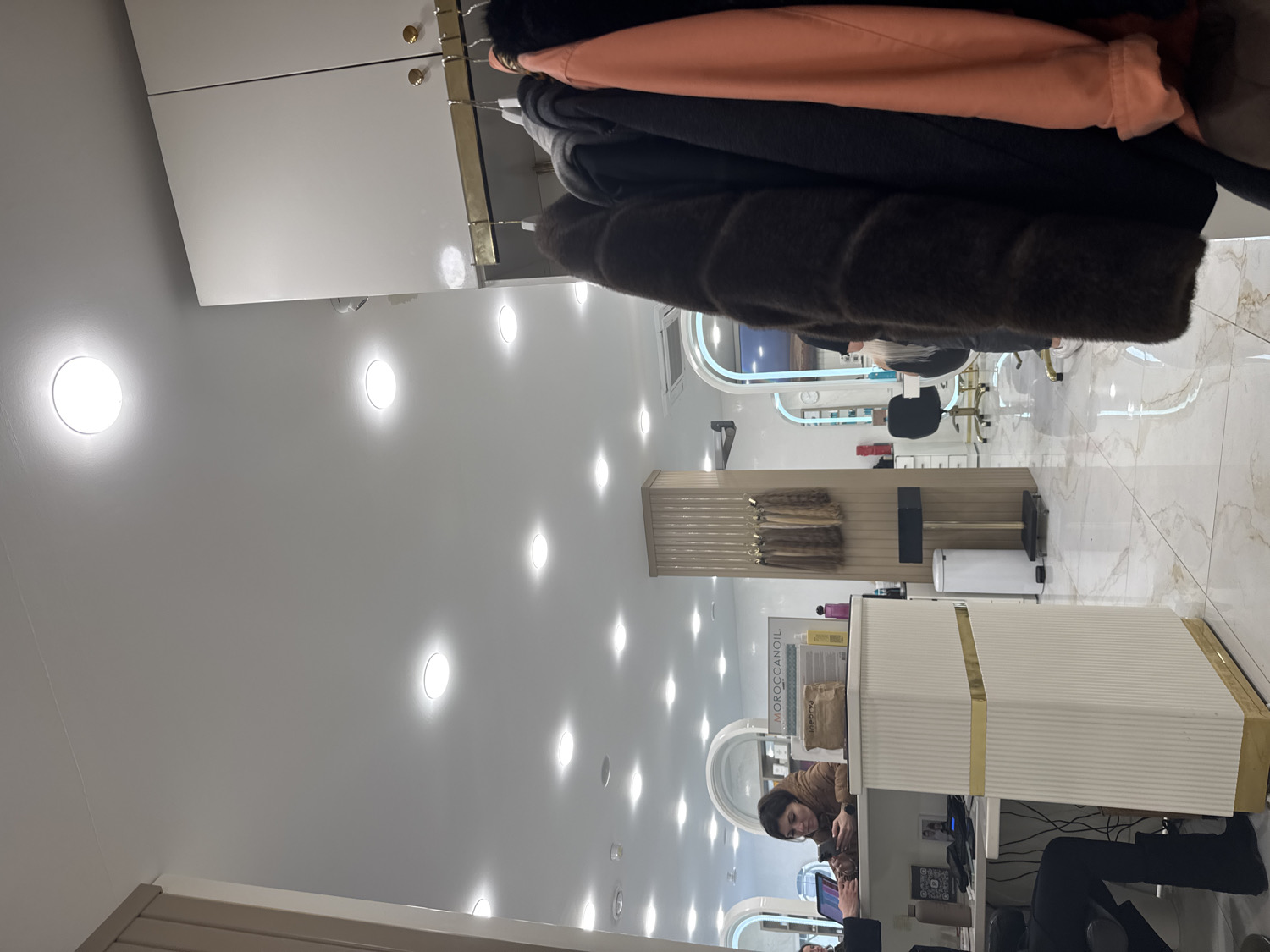 The image depicts the interior of a brightly lit hair salon or beauty studio. The white ceiling is illuminated by numerous circular, recessed lights, casting bright spots across the surface. The floor is covered with reflective, light-colored marble-patterned tiles.
In the upper foreground, a row of garments, including a peach-colored top, a black jacket, and a dark brown, possibly faux fur, coat, hang from an overhead rack. Above these, white cabinets with small gold handles are mounted.
To the right, a stylish reception or service counter, light-colored with gold accents and a ribbed texture, is prominent. Behind this counter, a woman with dark hair, wearing a brown jacket, is seated and looking down at a device she holds. Partially visible beneath the counter are the legs of another person, clad in black pants and boots. On the counter, various hair care products are displayed, including a bottle clearly labeled "MOROCCANOIL."
To the left of the counter, a tall display unit showcases several bundles of what appear to be hair extensions or hair swatches. Further into the background, multiple styling stations are visible, each featuring a gold-framed chair and a large arched mirror. Reflections in the mirrors hint at additional elements within the salon. A white cylindrical trash bin stands near the display unit.
The scene suggests a typical business day at the salon, with staff present and the space prepared for clients. The strong artificial lighting indicates it could be any time of day.