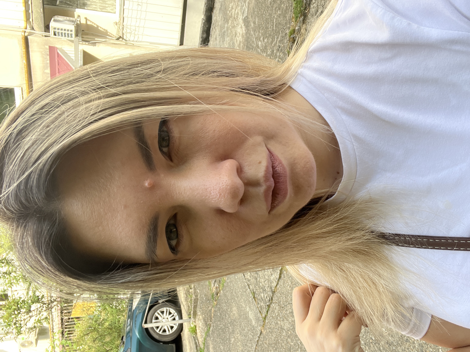 An outdoor, medium shot selfie captures a Caucasian woman with blonde hair, fair skin, and light green eyes. She is wearing a plain white t-shirt and a dark strap from a bag is visible across her chest. The woman has a small pimple on her forehead. Her left hand is raised and resting on her shoulder, with her fingers gently touching her hair.
The background suggests an urban environment. A dark green car with a silver wheel is parked on a paved surface. In the distance, a building with a light yellow facade and visible air conditioning units is present. The ground is concrete and somewhat uneven, with patches of grass. The overall lighting indicates it is daytime, with sunlight casting shadows. The specific location in Sochi, Russia, cannot be determined from the visual information alone.
