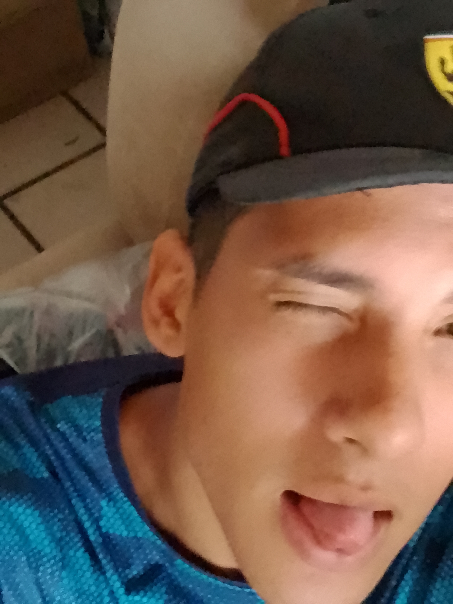 The image is a close-up selfie of a young person wearing a dark baseball cap with red trim and a yellow logo. The person is winking and sticking their tongue out slightly. They are wearing a blue t-shirt with a geometric pattern. The background appears to be indoors, with a tiled floor and some blurred objects. The lighting suggests it might be daytime. The overall impression is of a casual and playful moment. There is no information in the image to suggest it is located in La Guardia, Venezuela.