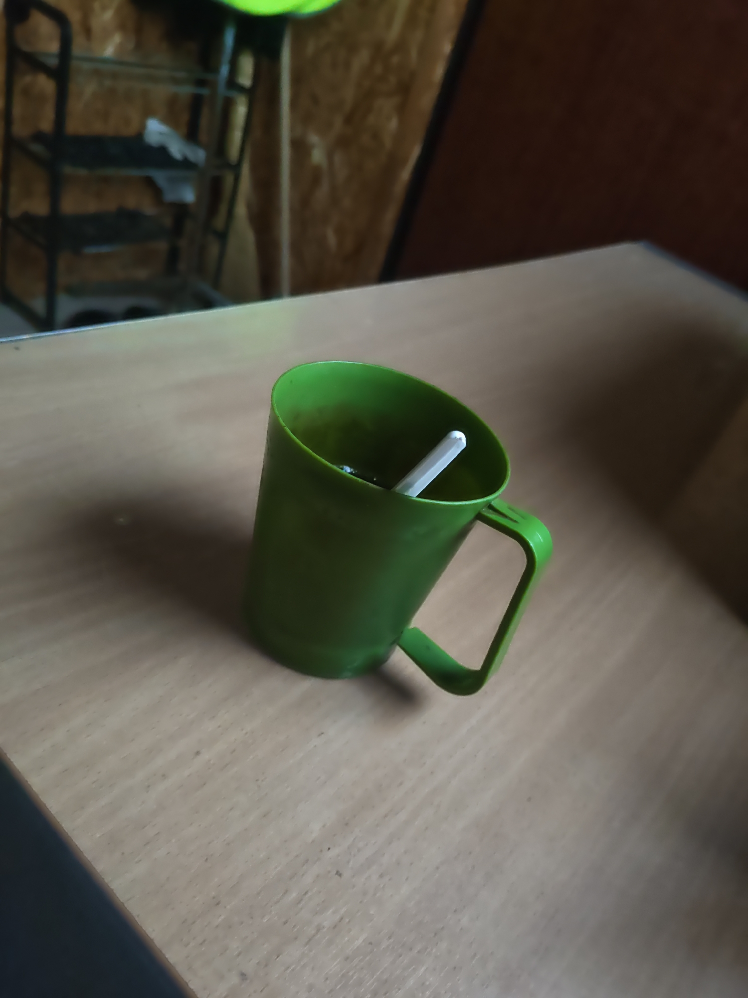A tilted, green plastic cup sits on a light-colored wooden table. Inside the cup, a white object, possibly a straw or utensil, is visible. The background consists of a wooden wall and a metal shelf unit. The scene appears to be indoors, possibly in a rustic or storage area. There is no indication of the location being Platoshino, Russia.