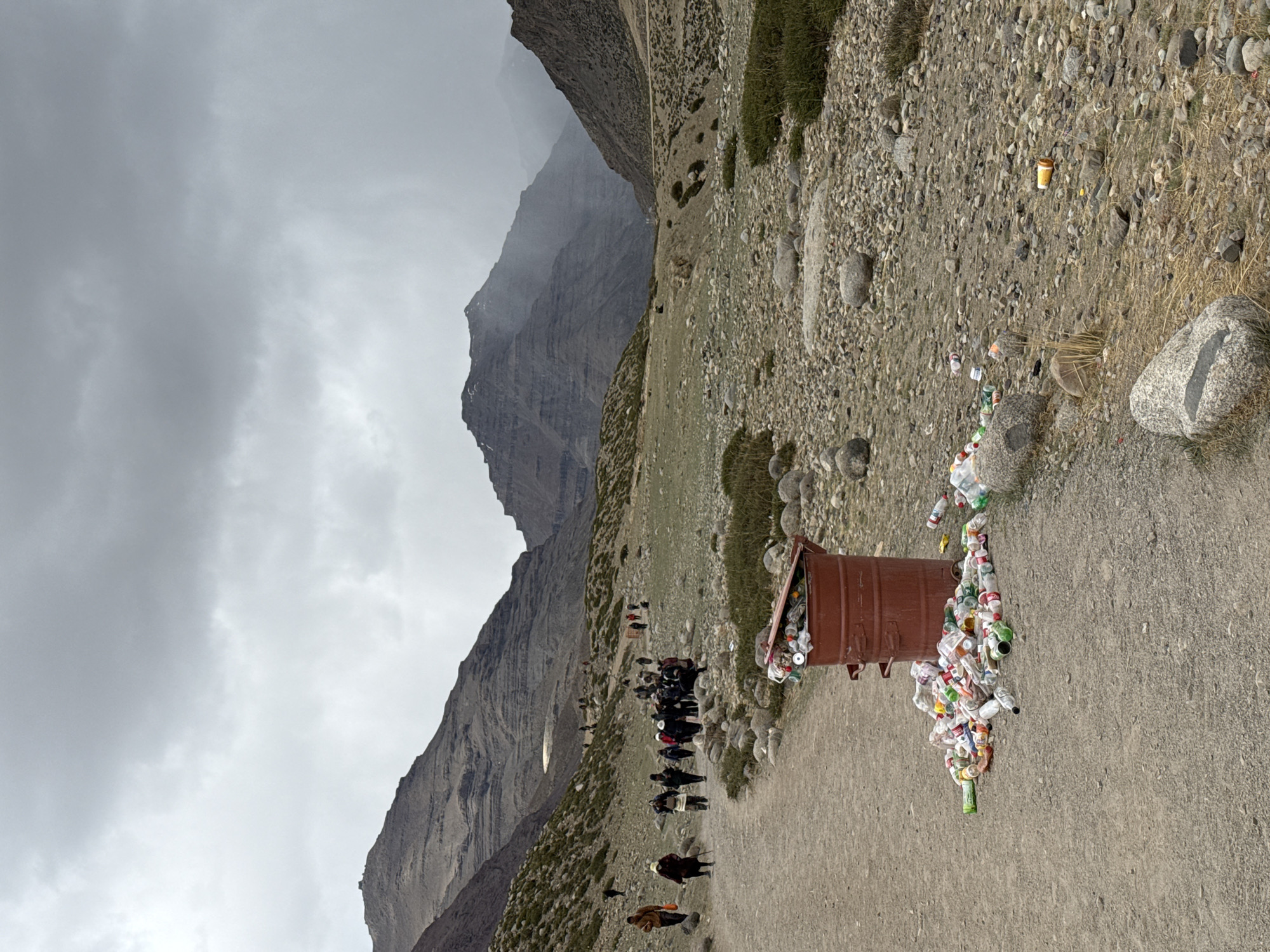 The image captures a rugged mountain landscape under an overcast sky, with a dirt path winding through the scene.
**Subjects:**
A long line of people, appearing to be trekkers or pilgrims, traverse the path from the midground towards the background. Many are walking, some carrying walking sticks, while others are riding horses or mules. Pack animals are also visible, suggesting a journey over difficult terrain. In the foreground to the right, a brown, cylindrical trash bin is heavily overflowing, with numerous plastic bottles, wrappers, and other refuse spilling out onto the ground. Additional litter, including cups and bottles, is scattered along the path in the foreground. The natural environment features towering, steep mountains in the background, their upper parts partially obscured by clouds or mist. The immediate terrain is rocky and arid, with sparse patches of grass and low-lying vegetation.
**Activity or event taking place:**
The primary activity is travel or trekking along a mountain trail. The continuous line of people and animals suggests a well-used route, possibly a pilgrimage or a popular hiking path, requiring significant effort given the mountainous environment. The overflowing trash bin indicates a gathering point or a frequently visited spot along the trail.
**Notable details:**
The time of day is daytime, but the sky is completely covered by heavy, grey clouds, creating a somber and diffused light. The weather appears cold and possibly damp. The mountainous landscape is stark and impressive, characteristic of high-altitude regions. The starkest detail is the severe accumulation of garbage around the bin, creating a significant visual contrast with the otherwise natural mountain environment. There is no discernible text visible in the image. While the provided context mentions "Parga town, China," the visual evidence (rugged Himalayas-like mountains, people's attire, pack animals) suggests a region more akin to the Himalayan ranges of India or Nepal, rather than a generic "Parga town."