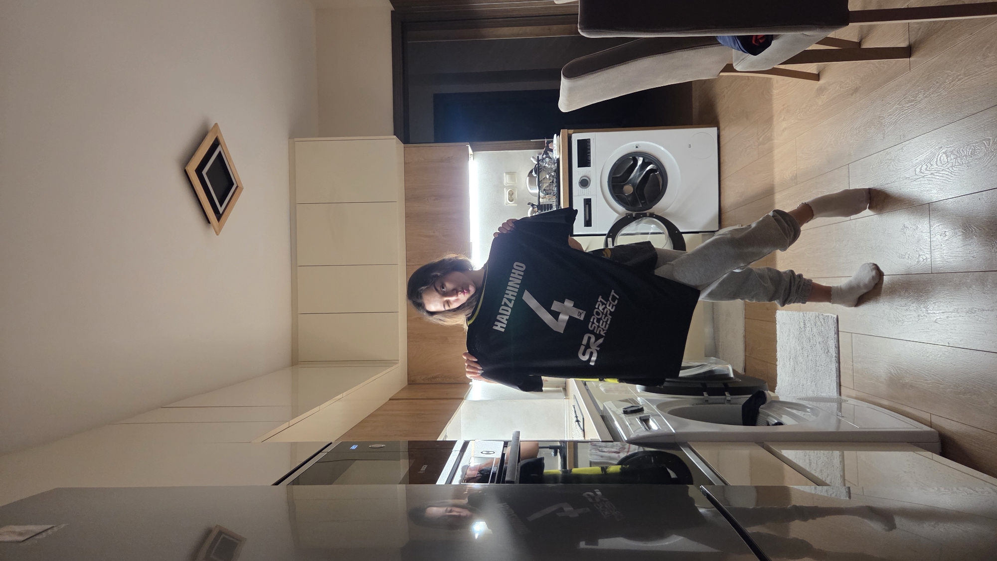A person in gray sweatpants and socks stands inside a modern kitchen. They are holding up a black soccer jersey with the name "HADZHINHO" and the number "4" printed on the back, along with the logo "SR SPORT RESPECT". The kitchen features light-colored cabinets, a washing machine, and stainless steel appliances. The environment appears to be a well-lit apartment or house. Location details (Stara Zagora, Bulgaria) are not visually discernible.