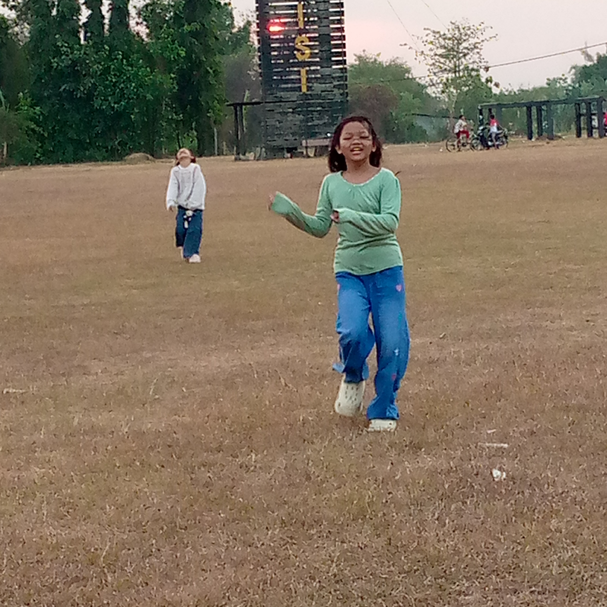 In Pacing, Indonesia, two young girls are playing in a dry, grassy field. One girl, wearing a light blue sweater and dark jeans, is running towards the right side of the frame, her head tilted back as if looking up. The other girl, in a green long-sleeved shirt and blue jeans with pink heart accents, is in the foreground, actively engaged in movement with her arms extended. She is wearing white Crocs. In the background, a tall wooden structure with horizontal bars and lights, possibly a scoreboard or a part of an obstacle course, stands prominently. Further in the distance, there are trees and some people on bicycles and a motorcycle. The overall scene suggests an outdoor recreational area.
