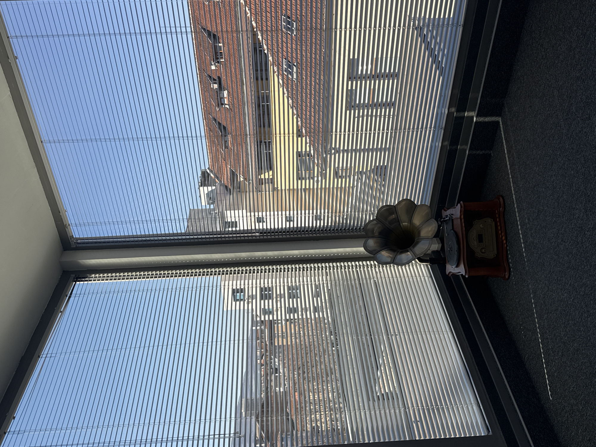 A view from inside a building in Sofia, Bulgaria, looking out through venetian blinds toward a city view. The blinds obscure the buildings outside, but glimpses of rooftops and buildings are visible. On the right side of the window, resting on the window sill, is a vintage gramophone. The foreground is a dark grey carpet.