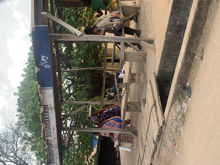 This image depicts a roadside scene in Egbedi, Nigeria, focusing on a rudimentary open-sided shelter identified as a "TRANSPORT ASSOCIATION MOTORCYCLE BKENGBWE" station. The shelter features a corrugated metal roof supported by rough-hewn wooden posts and beams. A banner on the roof displays the aforementioned text and a white motorcycle silhouette on a blue background.
Inside the shelter, five individuals are present, seated or resting on simple wooden benches and tables. From left to right:
1. A person in an orange and blue patterned dress is seated on a bench, facing away. A white bag rests beside them.
2. Another person, wearing a purple patterned outfit and a purple headwrap with long reddish-brown hair, is also seated on a bench, facing away.
3. In the mid-ground, a less distinct figure appears to be reclining or draped over a bench. Clothes are visible, possibly drying or stored, over a wooden beam. A sign on the ground reads "UNITED UNION".
4. A fourth individual, wearing a light-colored patterned tunic, is seated with their back partially to the camera, a colorful patterned bag resting near them.
5. The fifth person, wearing a green shirt and yellow trousers, sits further right, facing partially away from the camera.
The ground around and within the shelter is unpaved, appearing as dirt or sand. A concrete drainage ditch runs horizontally across the foreground, separating the shelter from the viewer. A makeshift wooden bridge, constructed from planks, spans this ditch, providing access to the shelter. Scattered debris, including plastic bags, is visible on the unpaved ground adjacent to the ditch. The background features dense green foliage from trees, under an overcast sky.