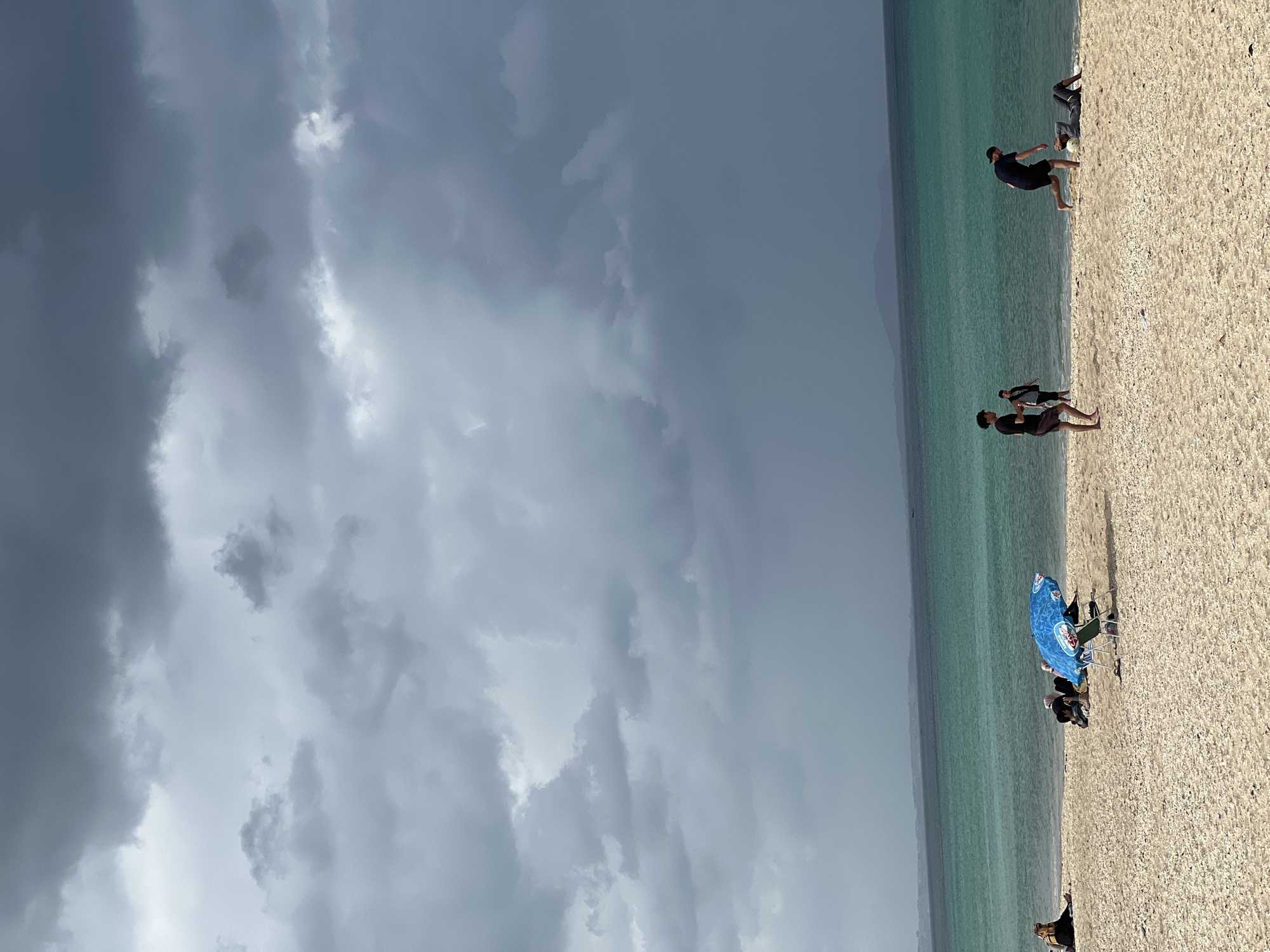 A sideways, medium-shot depicts a beach with people, water, and a stormy sky. The beach is composed of light-colored sand and what appears to be shells. Along the shoreline, the water is a clear turquoise. Further out, the water deepens to a slightly darker blue.
In the lower right corner, people sit under a blue beach umbrella with a white logo. Slightly up the shoreline are two people; one seems to be taking a photo of the other. Further up the beach are two men, one seemingly kicking a ball.
The sky is dominated by dark, ominous storm clouds.
Given the presence of the beach and the color of the water, the scene could plausibly be in M'Diq, Morocco. However, without additional landmarks or specific details, it's impossible to confirm the exact location.