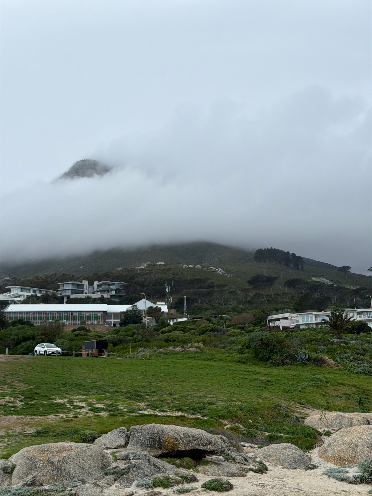 The image captures a cloudy day with a mountain peak shrouded in mist in the background. In the midground, several modern buildings with large windows are nestled amongst lush green vegetation and trees. One building appears to be a school or institution with a long, glass-fronted facade. A white SUV is parked on a grassy area near a small wooden structure. The foreground consists of a sandy beach with large, rounded rocks, some of which are covered in lichen. The scene suggests a coastal residential or institutional area in Cape Town, South Africa, possibly near a mountainous region like Lion's Head or Table Mountain, given the terrain and vegetation.