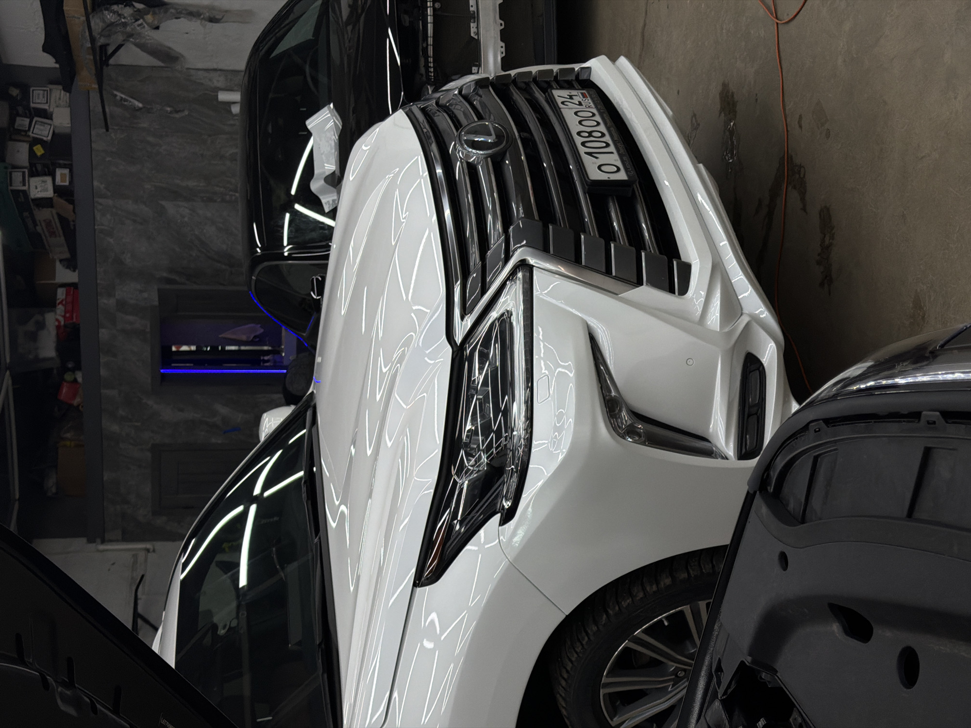 The image shows a white Lexus car with the license plate "O 108OO 24 RUS" in what appears to be an indoor garage or workshop. The presence of the license plate suggests that the car is likely located in Krasnoyarsk, Russia, as "24 RUS" is the regional code for the Krasnoyarsk Krai region. Other vehicles, also possibly undergoing repairs or detailing, are partially visible around the Lexus. The overall environment seems utilitarian and focused on automotive care.