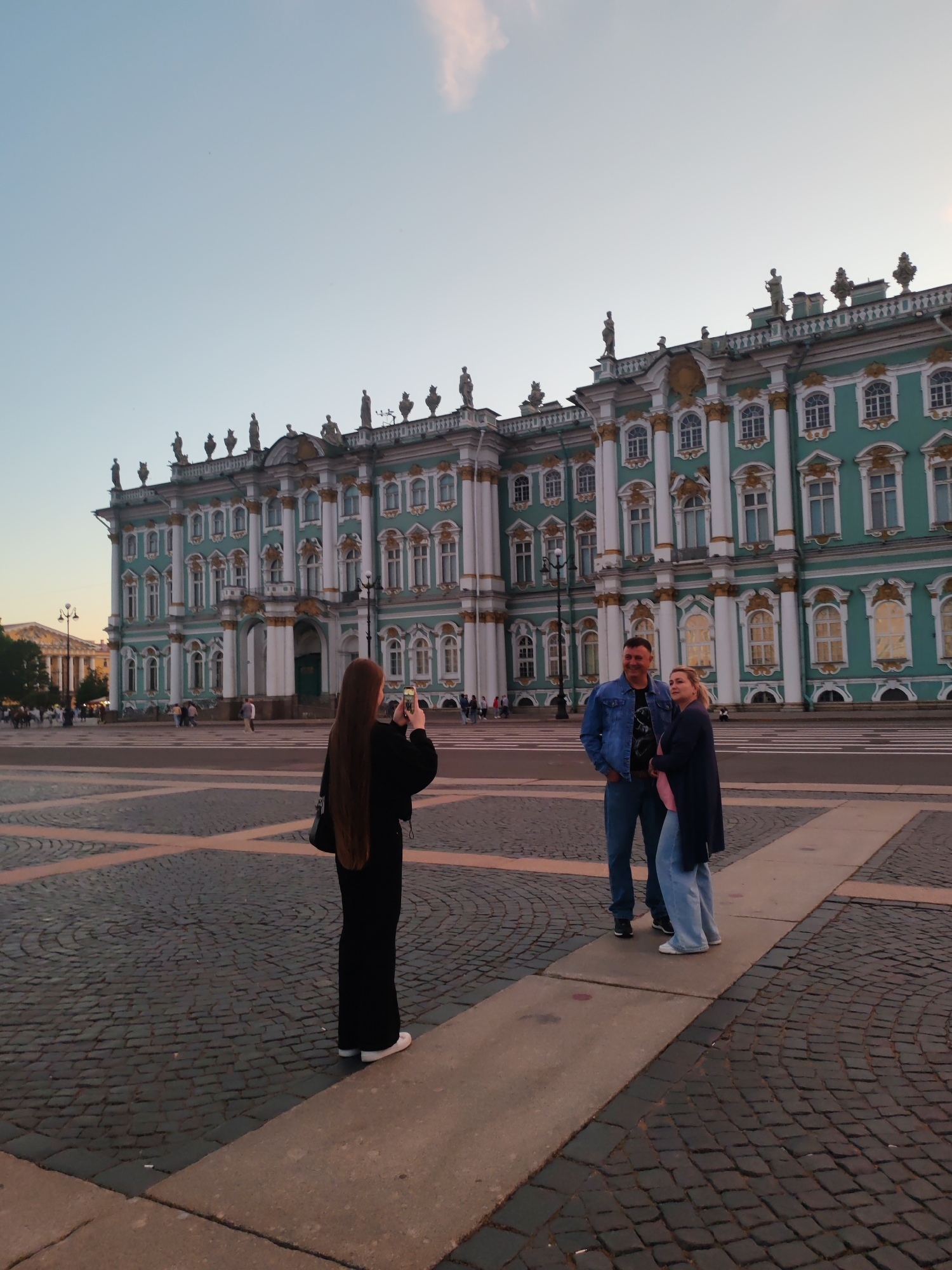 In Palace Square, Saint Petersburg, Russia, a woman with long brown hair is capturing a photo of a man and a woman standing together. The man is wearing a denim jacket and jeans, while the woman is dressed in a long cardigan and jeans. The Winter Palace, a grand Baroque building in a turquoise and gold color scheme, serves as the backdrop. The square itself is paved with cobblestones, and the sky above is a pale blue with a hint of clouds. The lighting suggests it is late afternoon or early evening, with the setting sun casting a warm glow. Other people are visible in the distance, and lampposts are sparsely placed throughout the square.