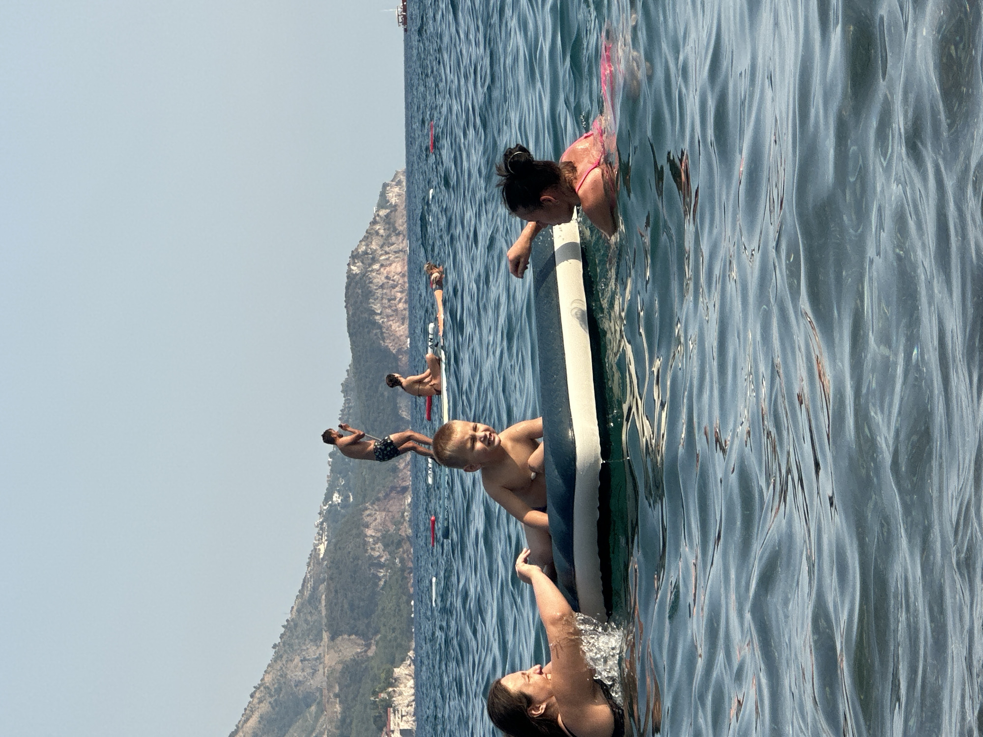 In Boreti, Montenegro, on a clear, sunny day, two women and a young boy are in the water. The boy is lying on an inflatable raft, his body from the waist up visible. A woman in a black swimsuit is to his left, her left arm extended towards him, splashing water. Another woman in a pink swimsuit is to the boy's right, her head and upper body visible as she looks towards the boy. In the background, a man in swim trunks stands on another inflatable raft, holding a phone. Further in the distance, a woman sits on an inflatable raft. The sea is blue and moderately wavy, with a mountainous coastline in the background. Several red buoys are visible in the water.