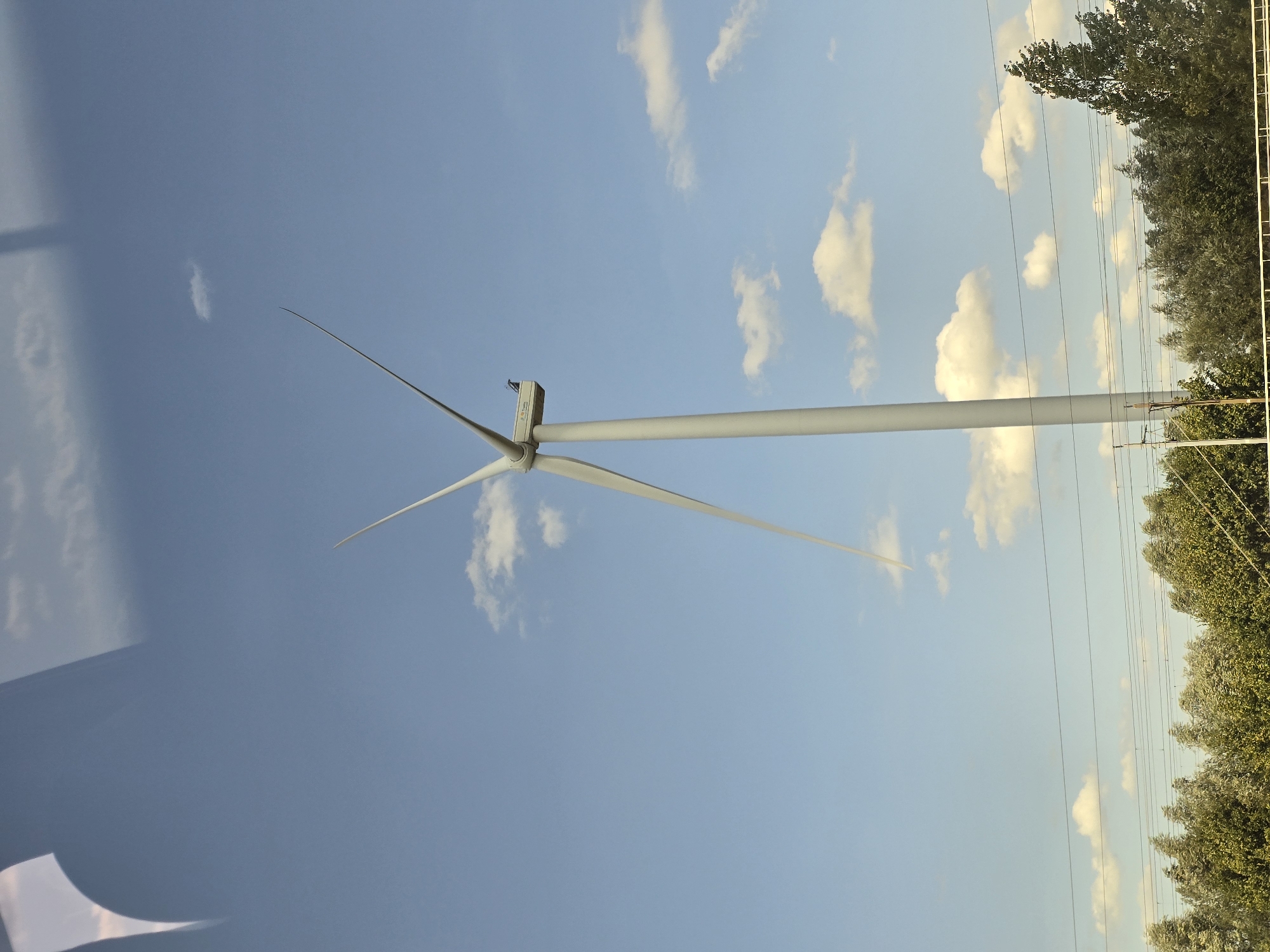 A lone, white, three-bladed wind turbine stands tall against a clear blue sky with scattered white clouds. The turbine is positioned in the lower right quadrant of the image. The lower portion of the image reveals a line of green trees, some of which appear wind-swept, suggesting a breezy day. Several horizontal power lines are strung above the trees, creating a network of dark lines against the background. In the upper left corner of the image, a blurred reflection of a window or surface is visible, overlaying the sky. The scene is tranquil and open, indicative of a rural or semi-rural setting.
