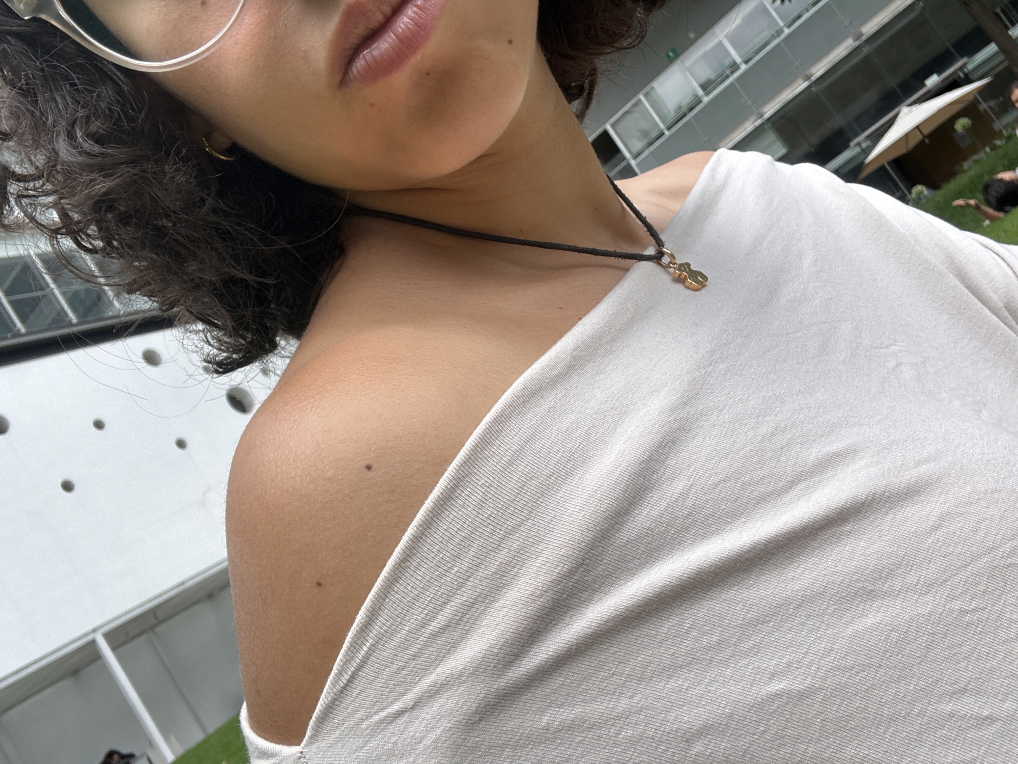 A close-up, eye-level shot depicts a light-skinned individual outdoors, with the top portion of their face visible, including clear eyeglasses and pursed lips. The person has dark, curly hair and is wearing a light-colored, off-the-shoulder shirt. A black cord necklace with a small, gold-colored pendant is visible around their neck. The left shoulder has at least two moles.
In the blurry background, a building with a white facade and multiple round indentations is partially visible. Additionally, there are glimpses of green lawn, a white parasol, and blurred figures of people in the background. The photo appears to be taken during daylight hours, suggesting an outdoor setting.
Based on the style of architecture, the prevalence of green space, and other contextual factors, this image was likely taken in Mexico City, Mexico. However, without further context, it's difficult to determine the exact location.