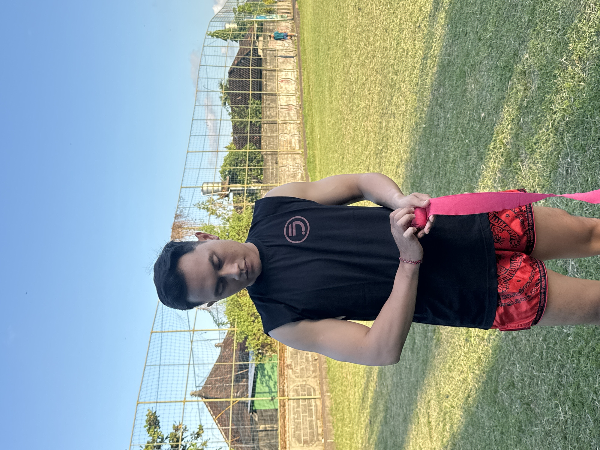 An adult male stands in a grassy field, wearing a black sleeveless t-shirt with a circular logo and red Muay Thai shorts with a tiger pattern. He is holding a pink hand wrap in his hands and appears to be in the process of wrapping it around his wrist. The background shows a chain-link fence with a tiled roof building behind it, a small water tower, and lush green trees, indicative of a tropical environment. In the distance, another person in a blue shirt can be seen walking across the field. The scene appears to be an outdoor sports or training area, possibly a local sports ground in North Kuta, Indonesia, given the architecture and vegetation. The lighting suggests it is late afternoon.