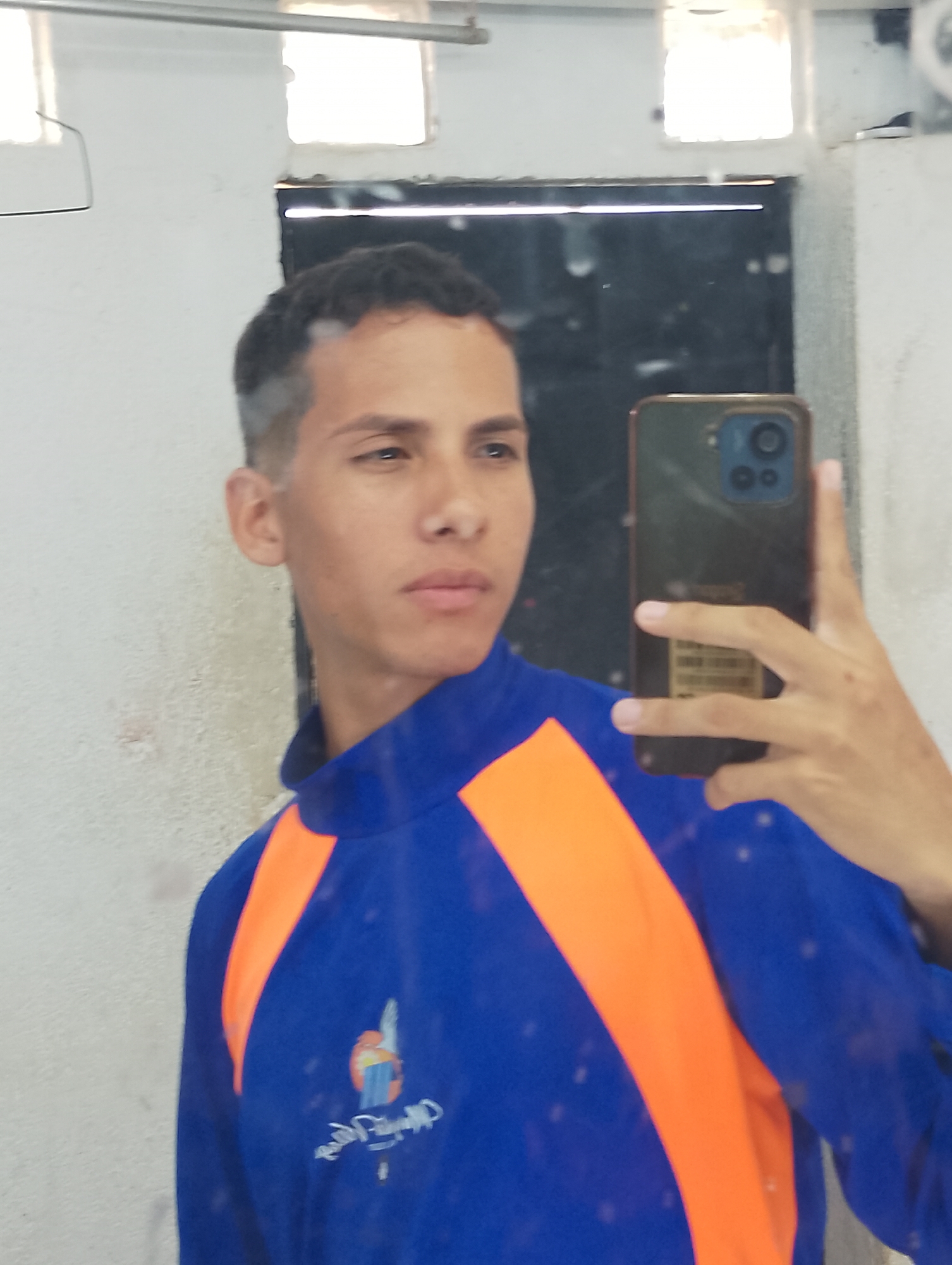A young man is taking a selfie in a bathroom mirror. He is wearing a blue shirt with orange stripes on the shoulders and a logo on the lower left side. He is holding a dark-colored smartphone with multiple cameras visible on the back. The bathroom appears to be indoors, with a dirty white wall, a metal rod above the mirror, and a rectangular ventilation window above the mirror. It is hard to determine the city in which this photo was taken. The picture would need to contain identifiers to provide the requested location information.