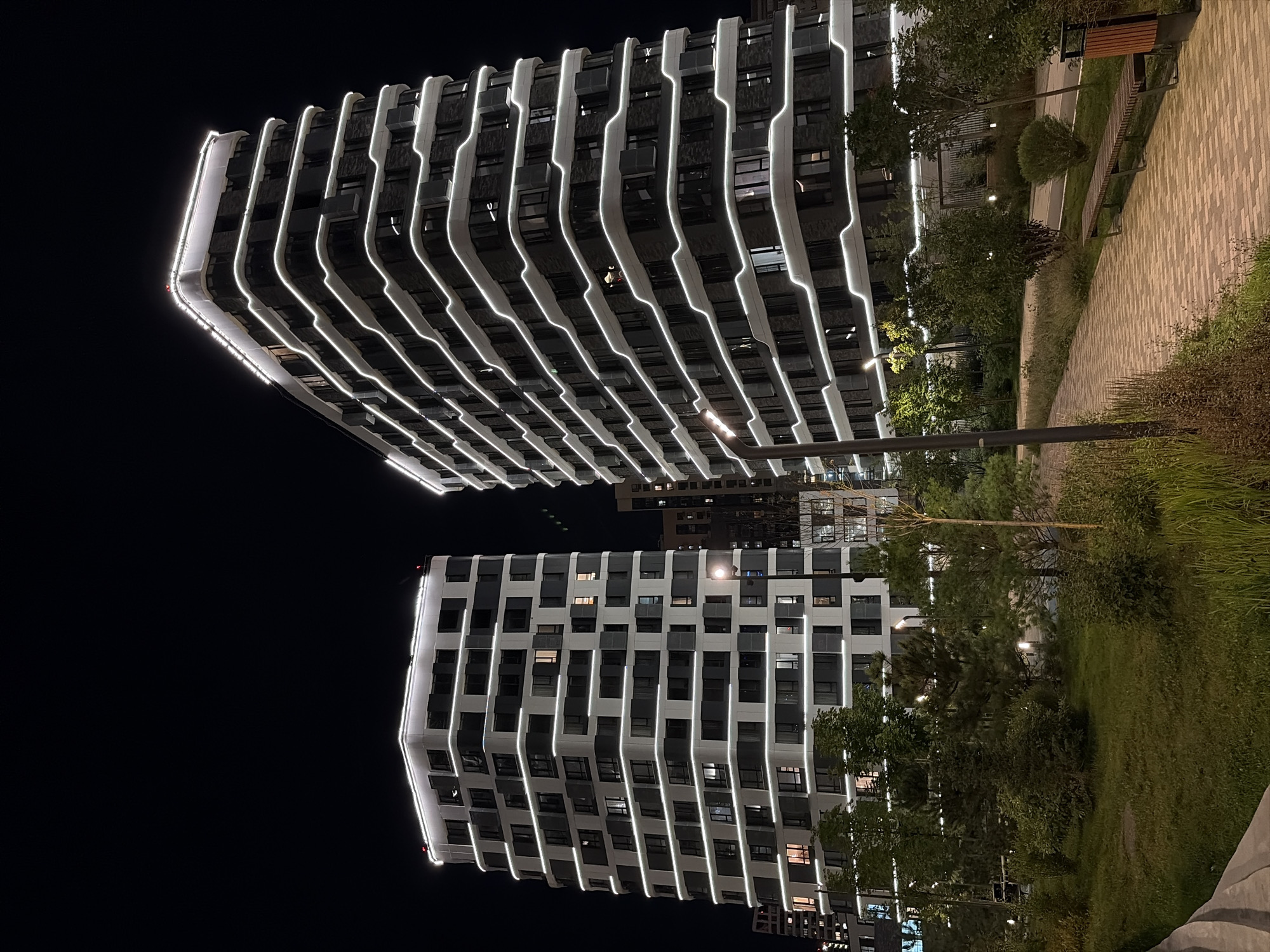 This is a nighttime exterior shot of two modern, multi-story apartment buildings in Moscow, Russia. The buildings are illuminated with white LED strip lights that highlight their architectural features, creating a striking contrast against the dark sky. One building, to the left, is squarer in shape, while the building to the right is taller and has a more curved facade. Both buildings feature numerous windows. In the foreground, a paved walkway with a herringbone pattern leads past a grassy area with trees and shrubs. A park bench and a trash receptacle are visible along the walkway. The overall scene suggests a well-maintained, contemporary urban residential area.
