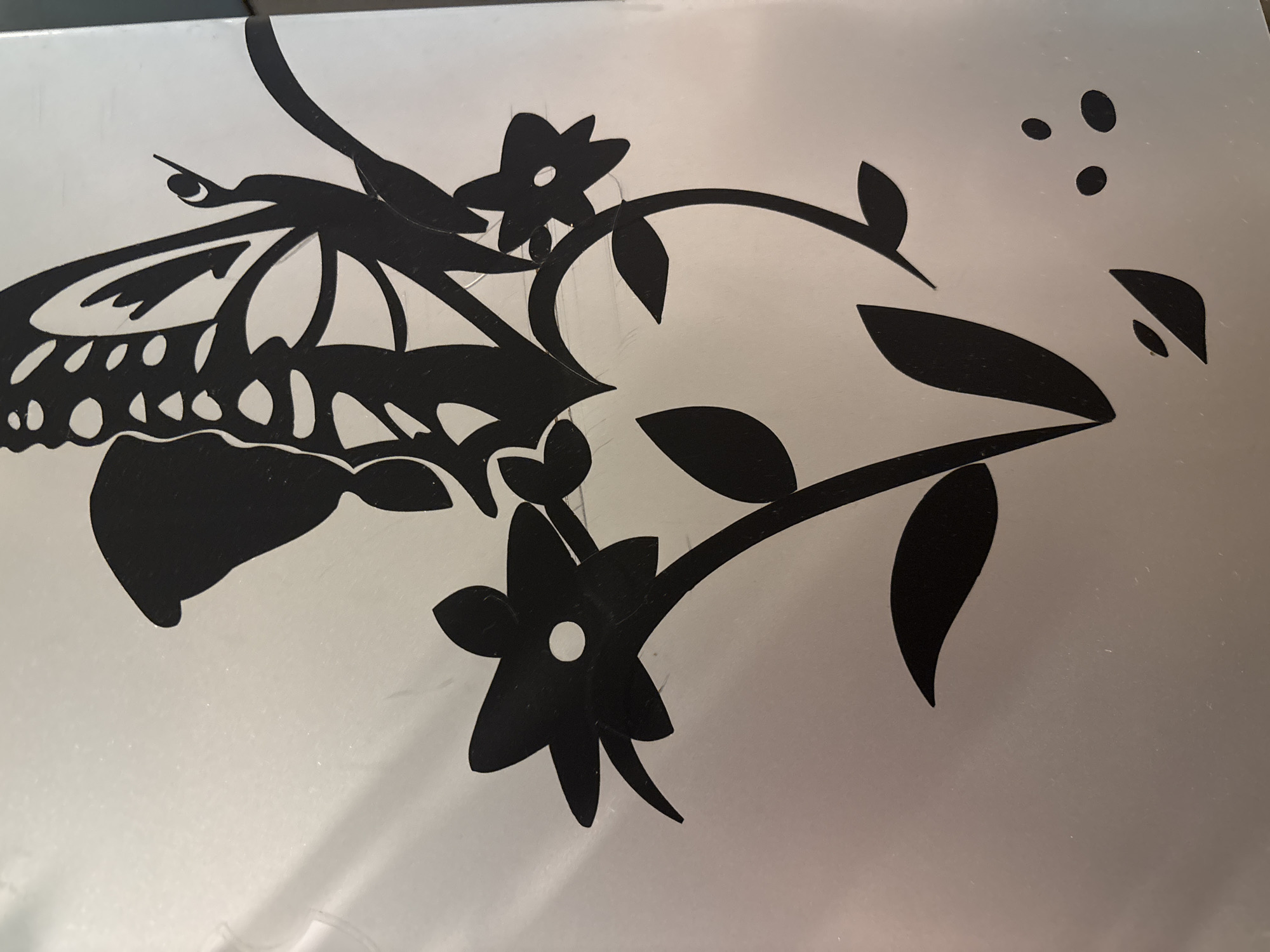 The media file presents a close-up view of a decorative design, rendered in black silhouette against a light gray, somewhat reflective background. The primary subjects are a stylized butterfly and a floral vine motif.
On the left side of the image, a large butterfly is depicted with intricate patterns forming its wings and a slender antenna extending upwards. Adjacent to the butterfly, a winding vine branches out, adorned with several leaves and flowers. Two prominent flowers are visible, each characterized by a small, uncolored circular cutout in its center, suggesting a staminal area. Further to the upper right, three small, solid black circular shapes are scattered, possibly representing berries or additional decorative elements.
The background surface exhibits subtle reflections and minor, faint scratch marks, indicating it may be a metallic or laminated material. The lighting is even, without harsh shadows. The image captures a static, decorative pattern, offering no discernible activity or event. There is no visible text within the frame. The object depicted is located in Vagharshapat, Armenia.