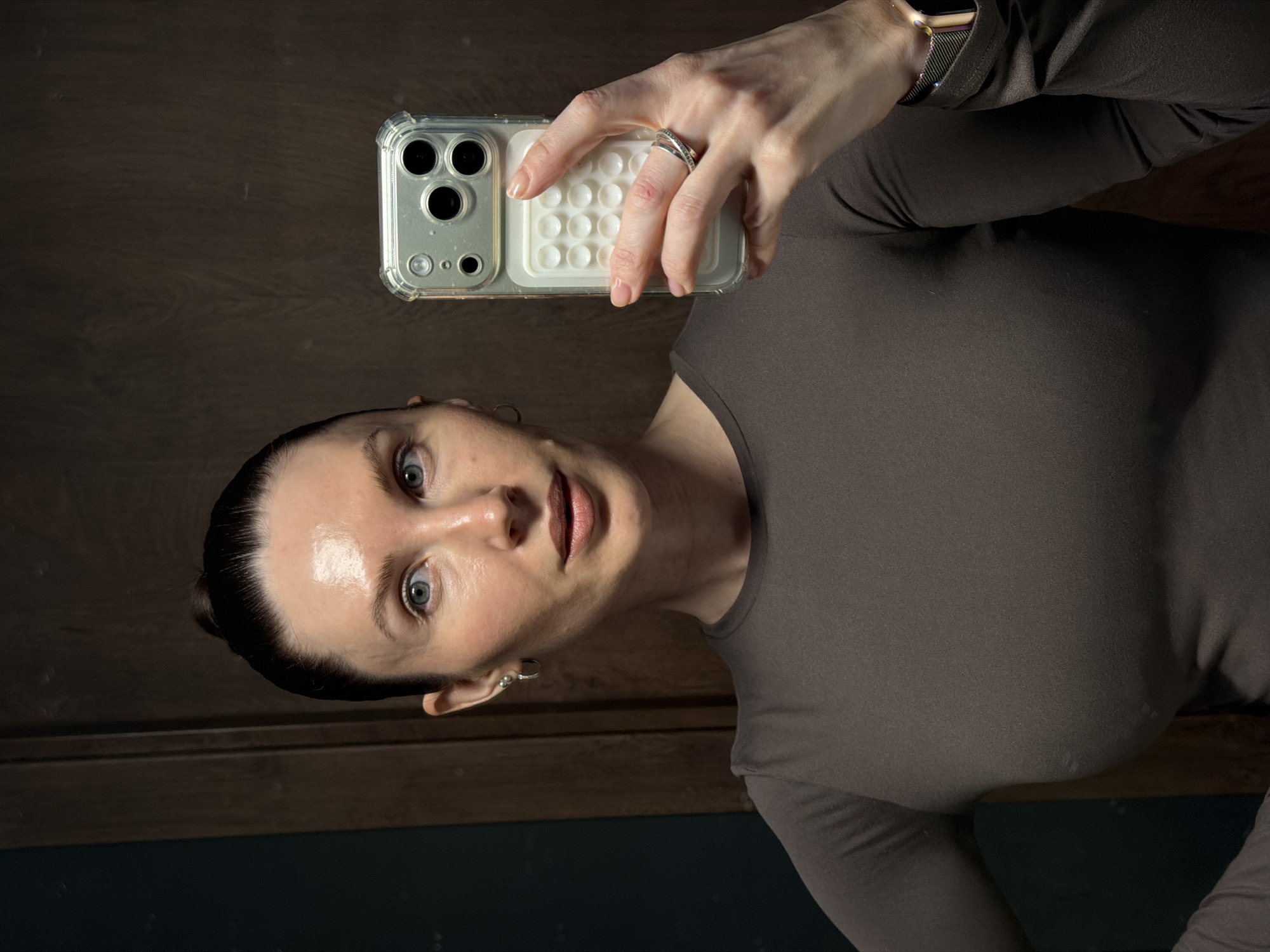 A woman is taking a selfie. She is wearing a dark brown long-sleeved shirt and a watch. Her phone has a clear case with a white pop-it style grip on the back. She is wearing a ring on her ring finger. The background appears to be a dark brown wall or door. The location is unknown and is not identified as Perm, Russia.