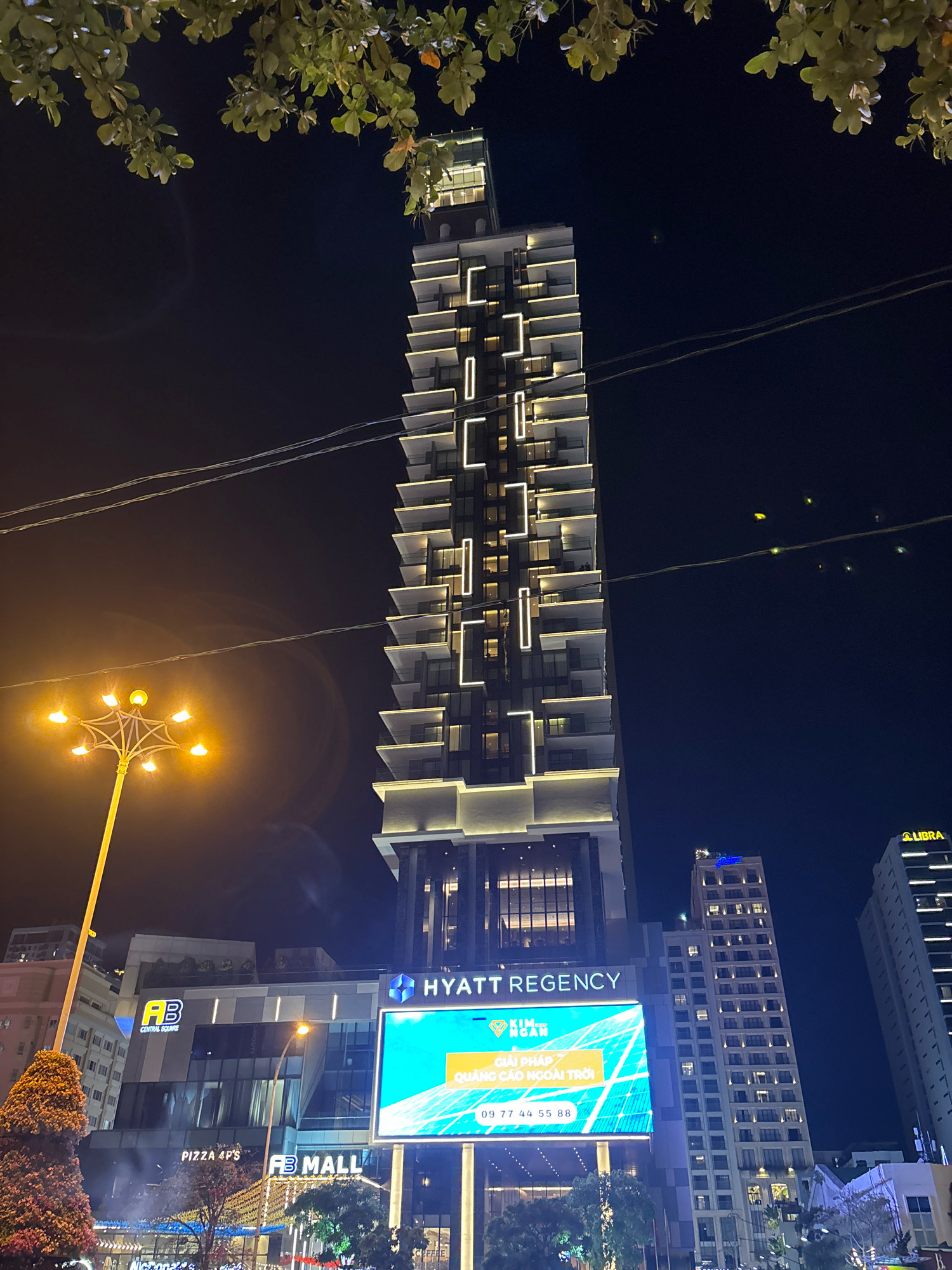 The image captures a nighttime scene in Nha Trang, Vietnam, dominated by the striking Hyatt Regency hotel, a tall, modern skyscraper illuminated with white lights tracing its balconies and architectural features. In front of the hotel, a large LED billboard displays vibrant graphics and text, including the words "HYATT REGENCY" and "KIM NGAN," along with a phone number. To the left, a prominent street lamp with multiple yellow bulbs illuminates the foreground, casting a warm glow. Adjacent to the Hyatt, other buildings are visible, including one with a "LIBRA" sign and another bearing "AB CENTRAL SQUARE." A tree with reddish foliage stands in the lower left, and the night sky above is dark, with a few faint lights or celestial objects visible. The overall impression is of a lively urban environment at night, with modern architecture and prominent signage.