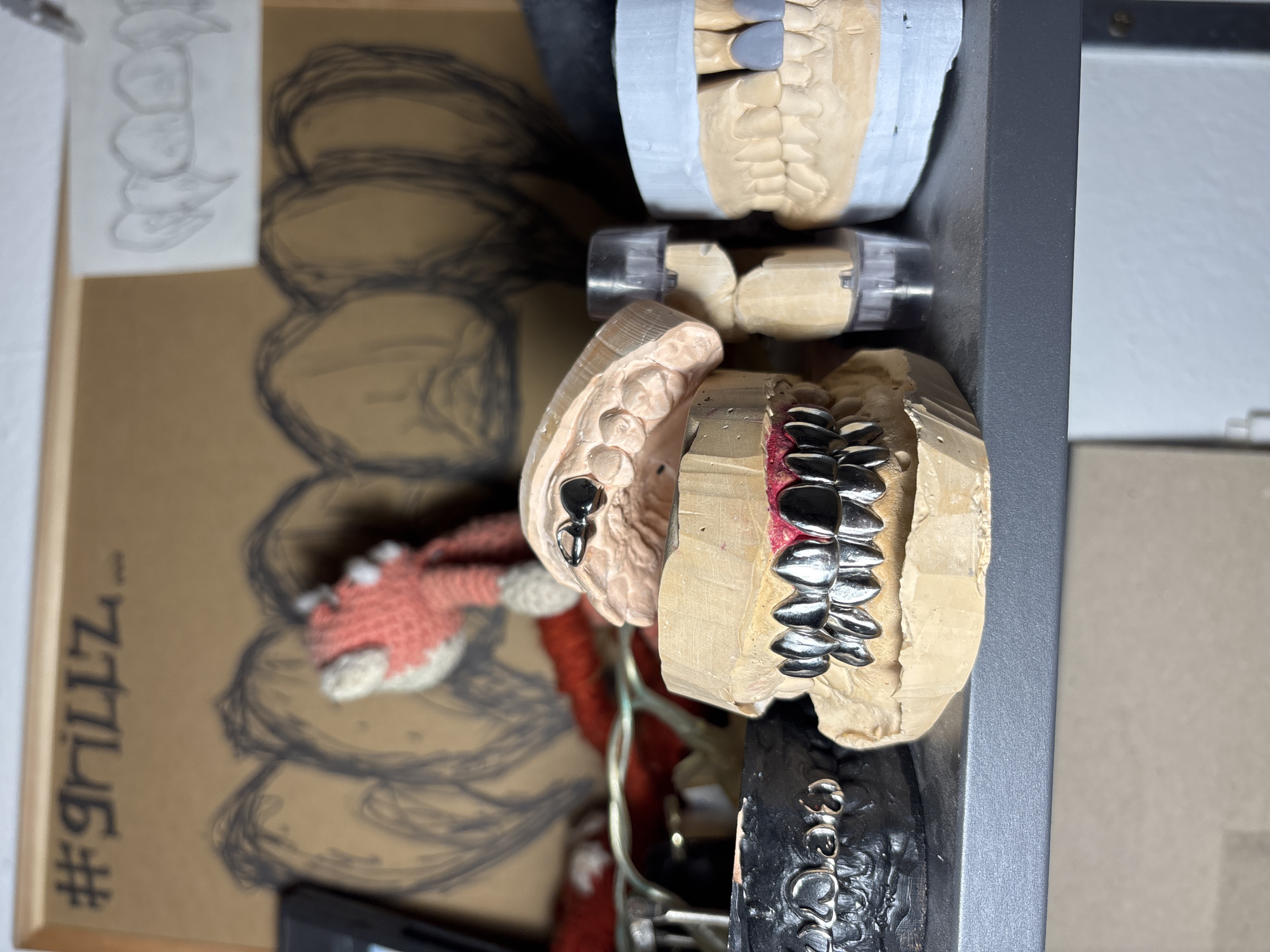 The image shows a cluttered shelf containing dental molds, likely for the creation of grillz. The molds are arranged on the shelf, with some having silver grillz fitted onto them. A drawing of teeth and a hashtag labeled "#grillzz" can be seen on a framed picture in the background. The location is unknown and there is no information regarding the city of Sofia, Bulgaria.